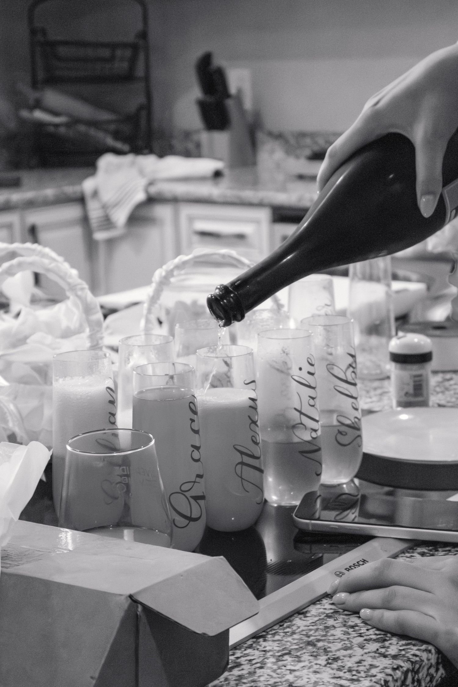 Champagne being poured into personalized bridesmaid glasses on a kitchen counter in black and white.