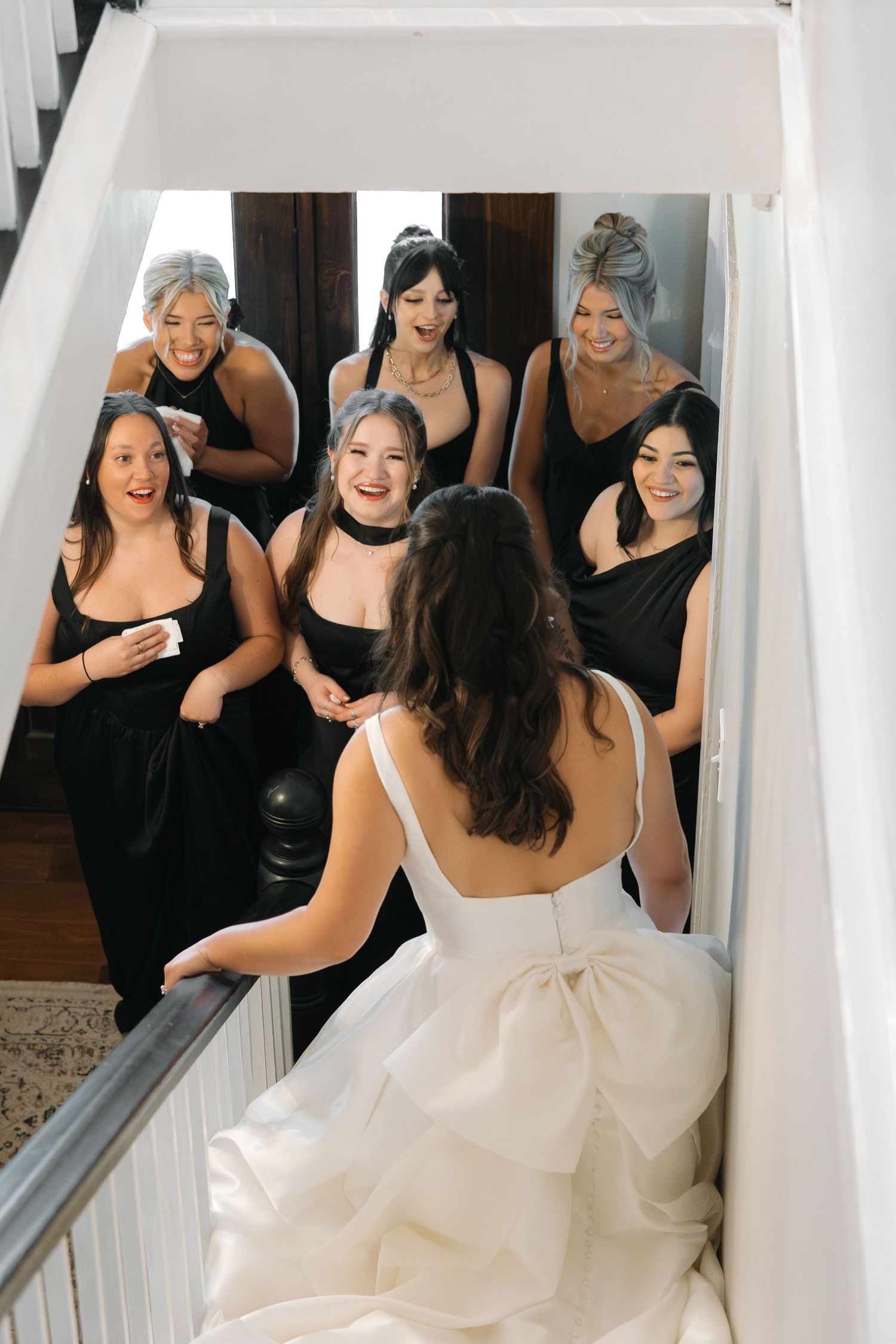 Excited bridesmaids in black dresses react with joy seeing bride in white gown on staircase.