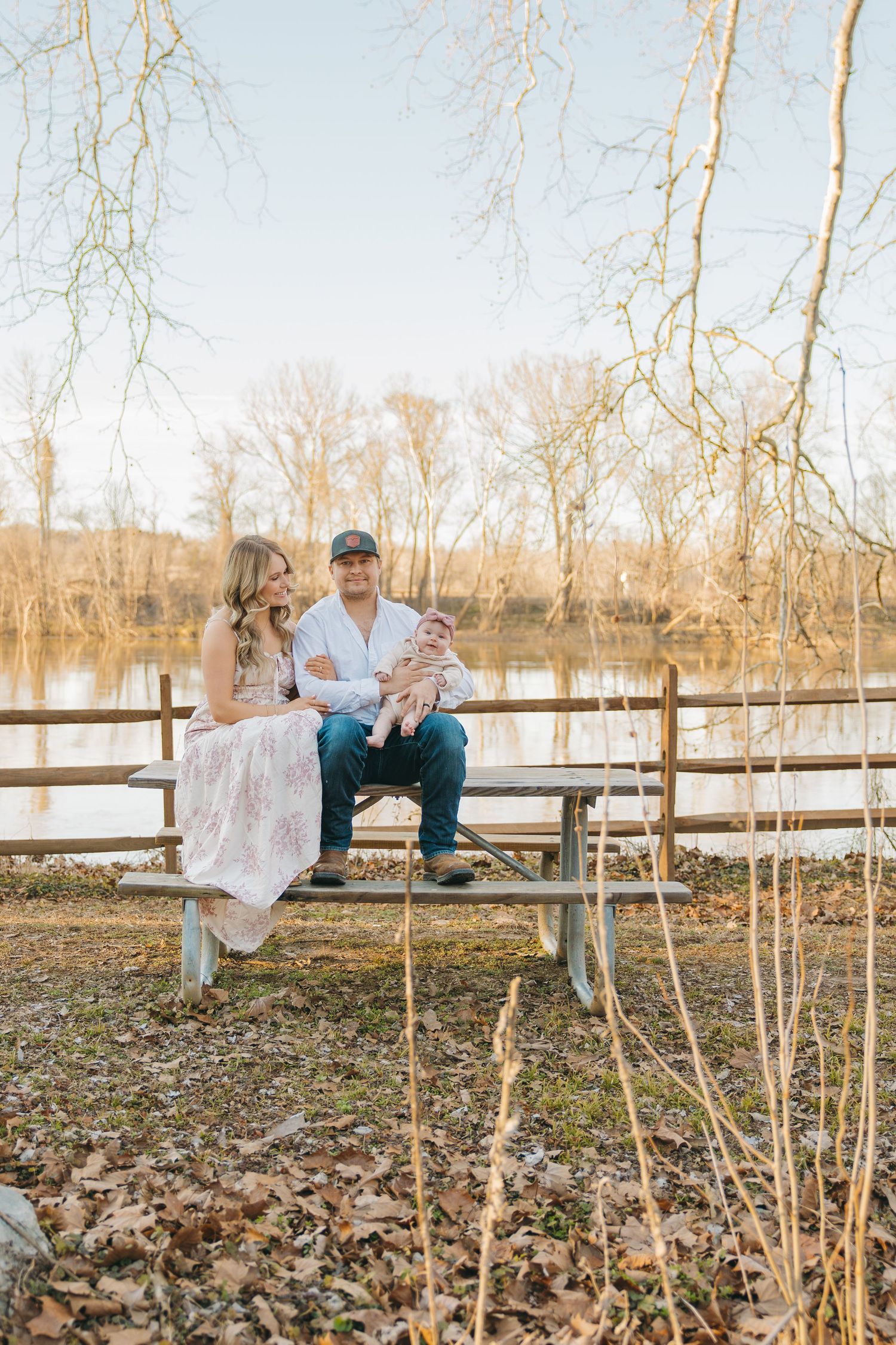 Couple sits on a wooden bench near a lake holding their newborn baby surrounded by bare winter trees.