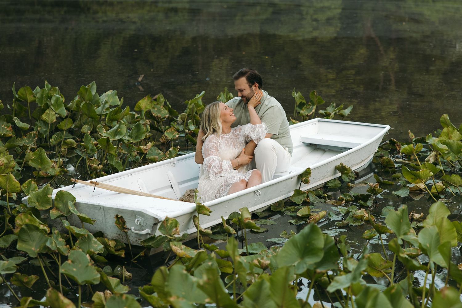 A couple shares an intimate moment kissing in a white rowboat nestled among lush green lily pads on a lake.