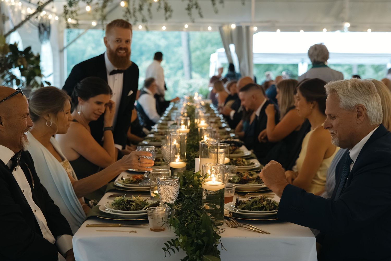 Elegant wedding reception dinner under tent with candlelit long tables and greenery centerpieces.