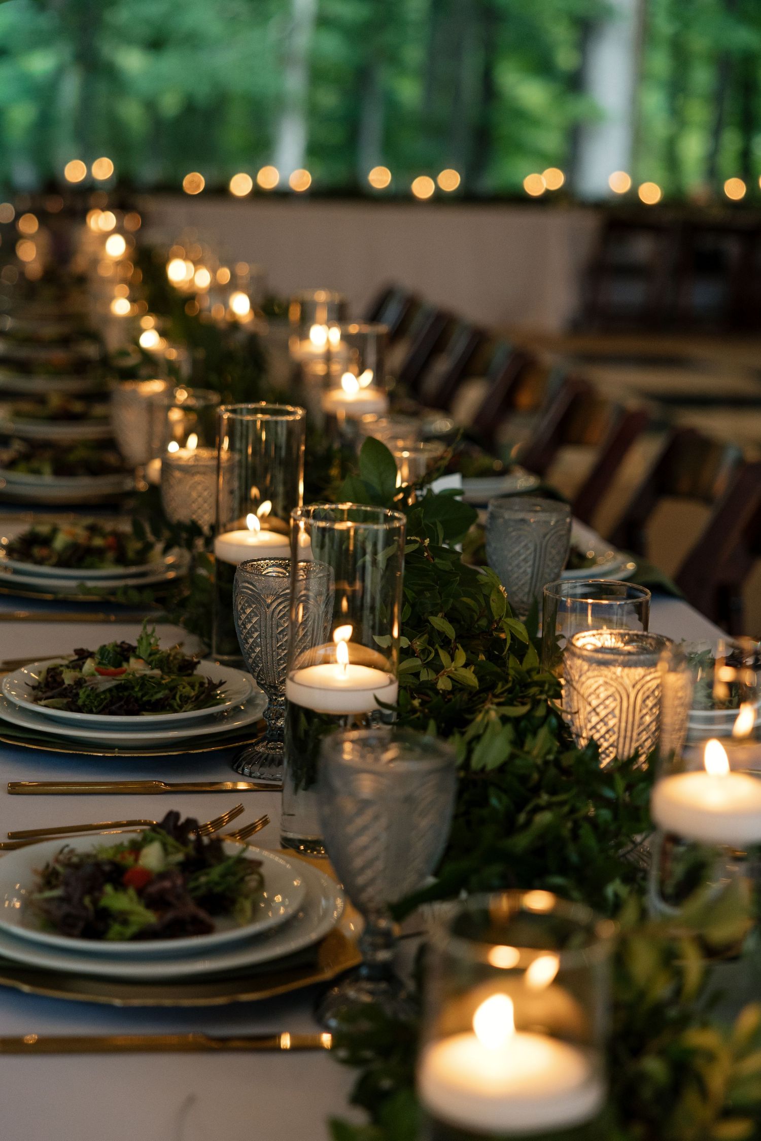 Elegant candlelit wedding reception table with greenery runner, crystal glasses, and gold cutlery.