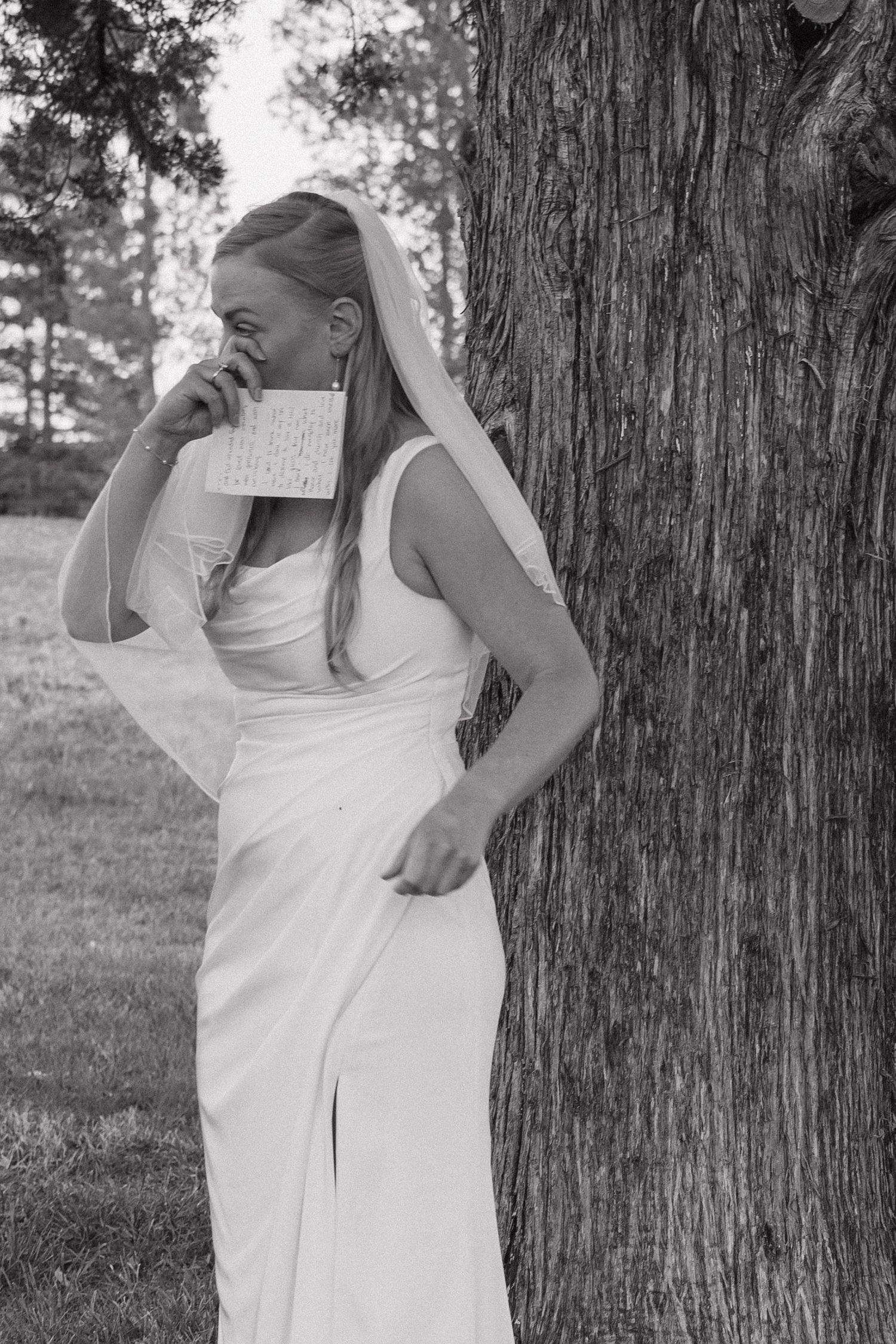 Emotional bride in white gown and veil reading a letter while leaning against a tree, black and white.