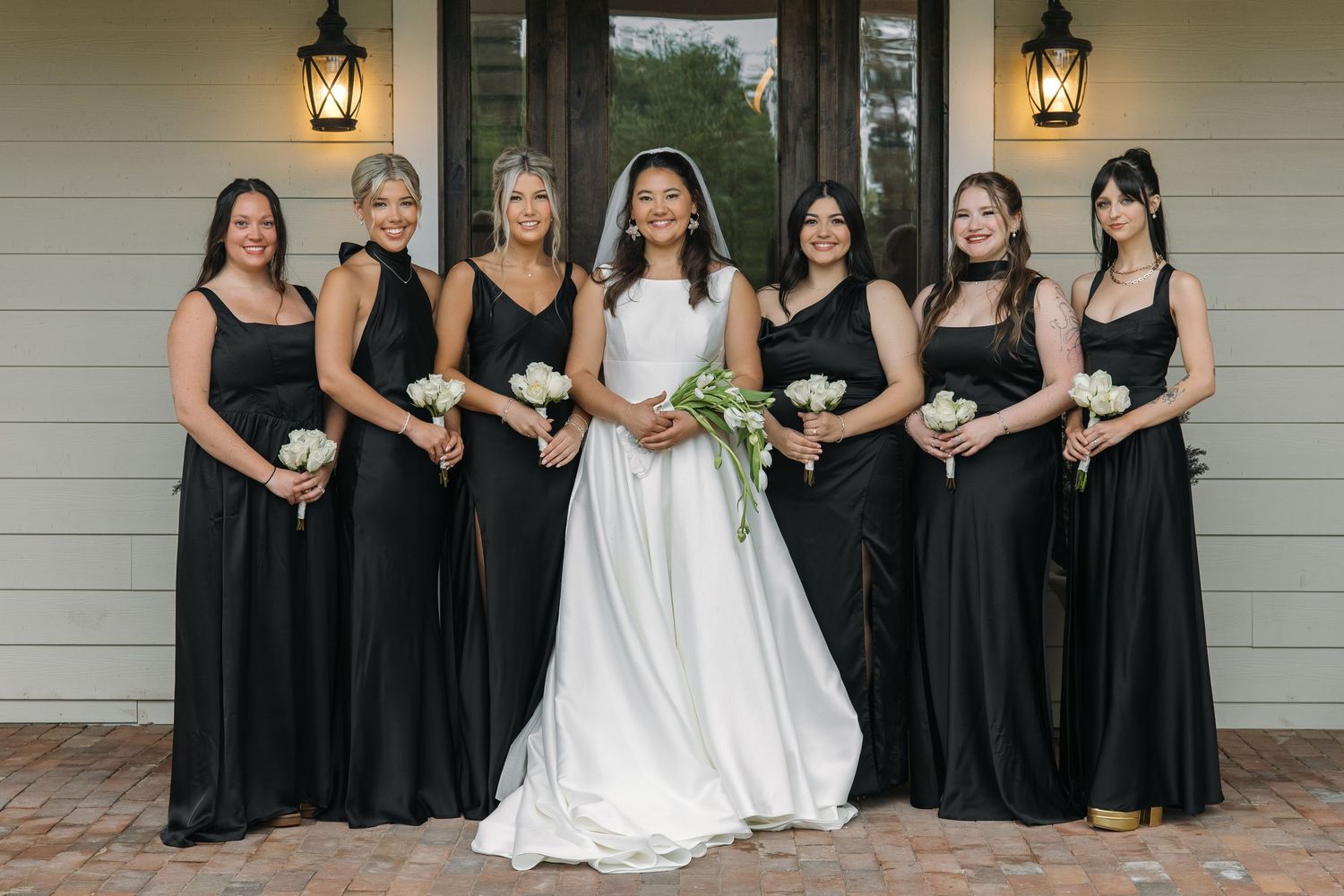 Bride in white gown poses with six bridesmaids in black dresses holding white bouquets outside elegant home.