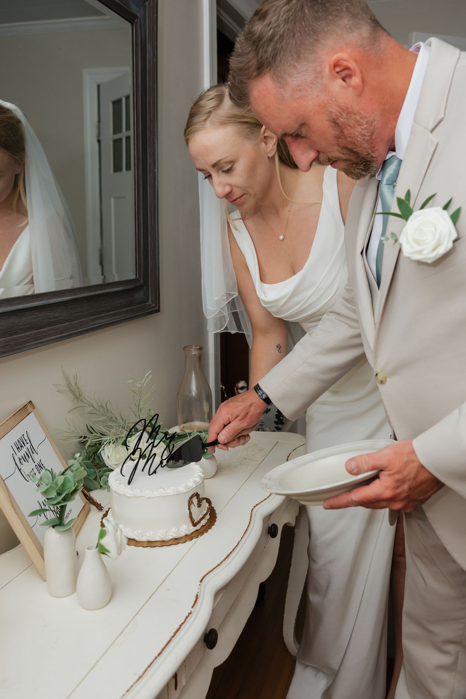 Bride and groom cut their white wedding cake decorated with greenery and calligraphy topper together.