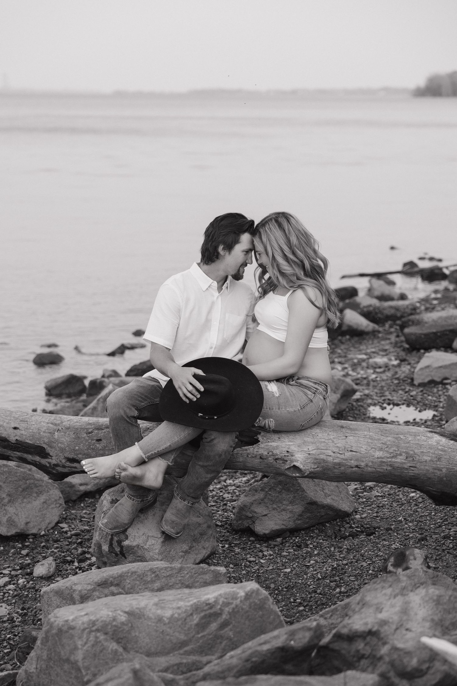 Black and white photo of couple sharing intimate moment on rocky beach, man holding cowboy hat near pregnant partner.