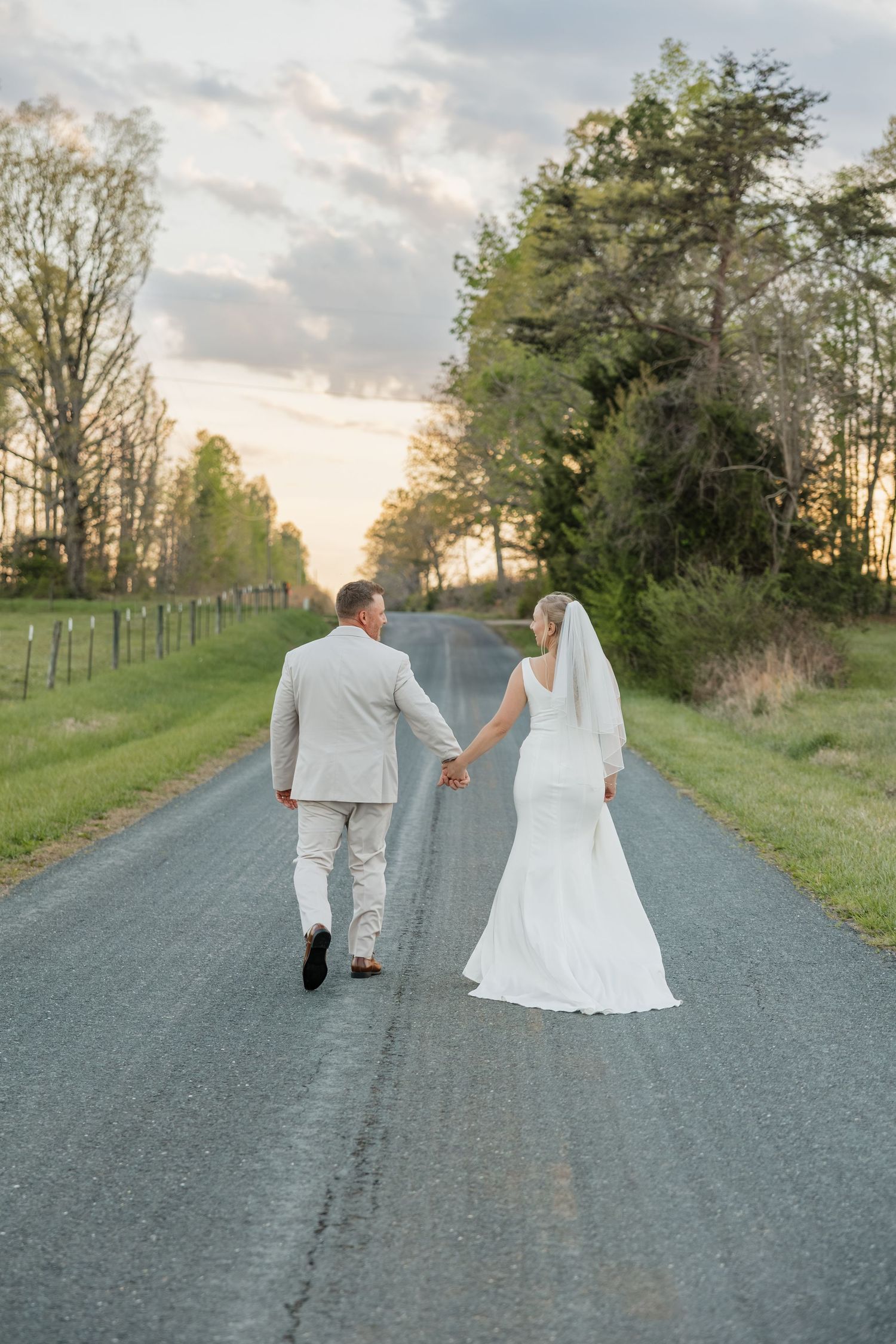 Bride and groom walk hand in hand down a quiet country road at sunset.
