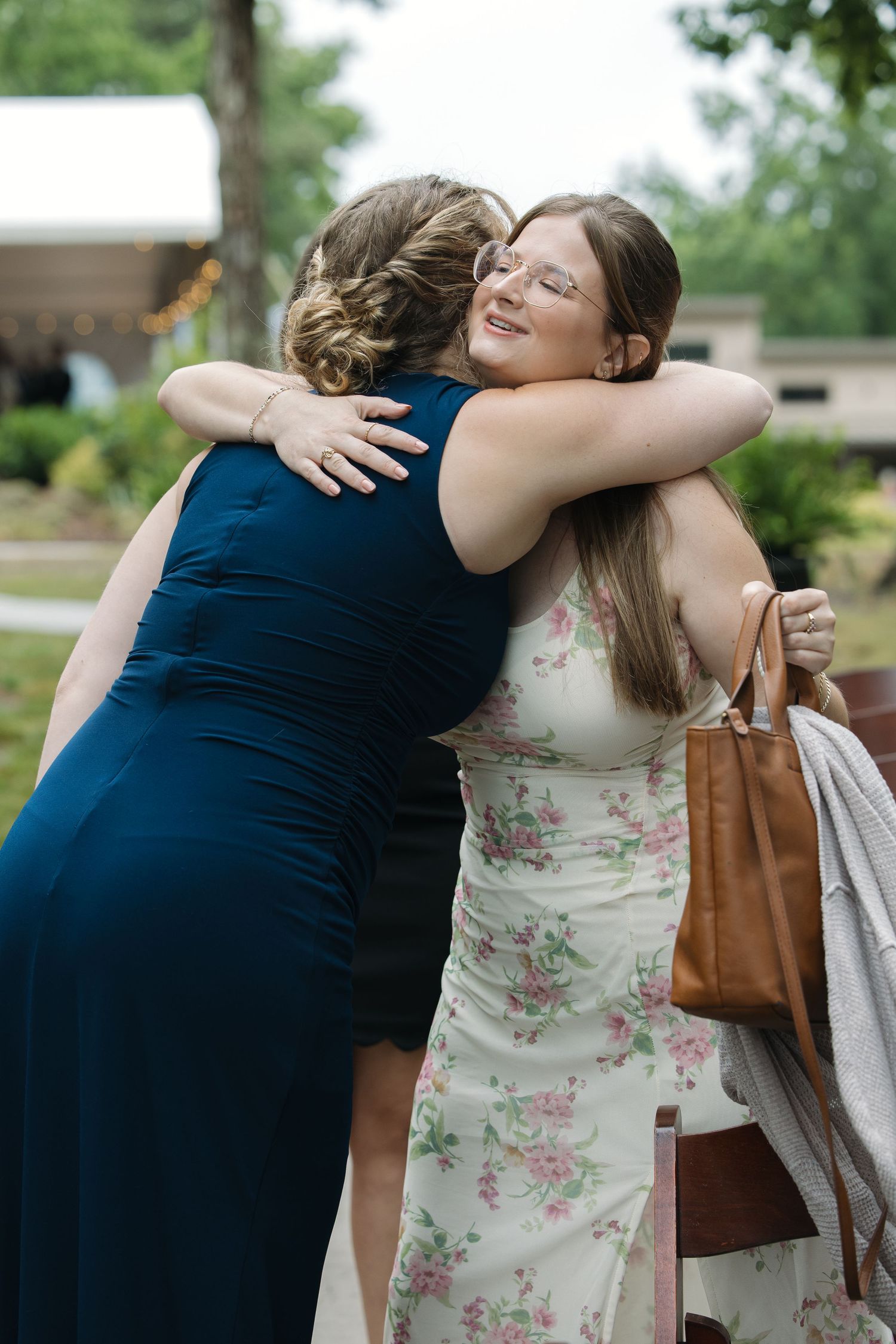 Two women share a warm embrace outdoors at a wedding celebration in summer.