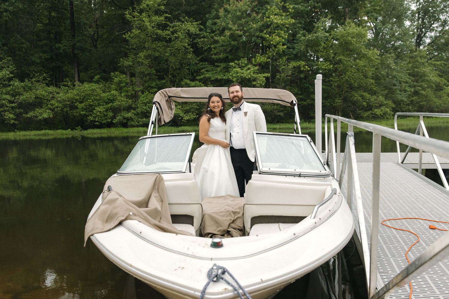 Newlywed couple in white wedding attire posing together on a motorboat docked at a lakeside pier.