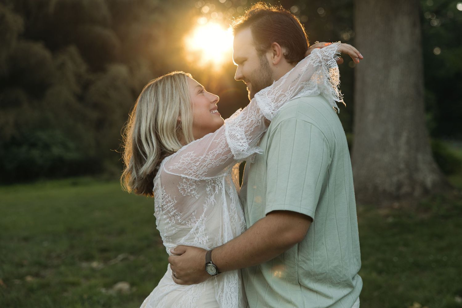 Couple gazing lovingly at each other during glowing sunset in lush green park, woman in white lace dress.