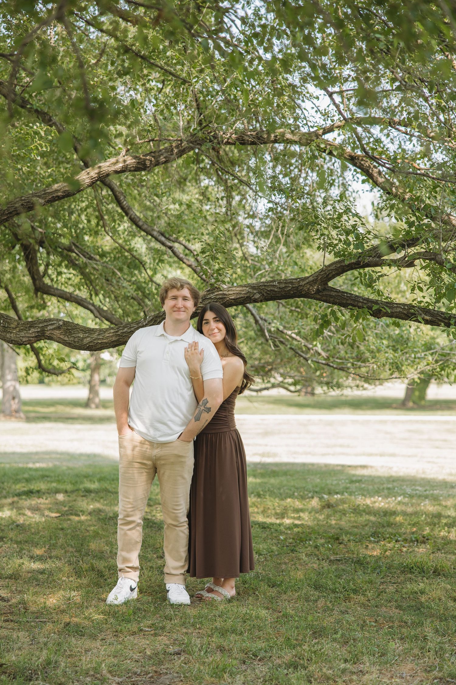 A couple stands embracing under a large tree in a lush green park, man in white polo and woman in brown dress.