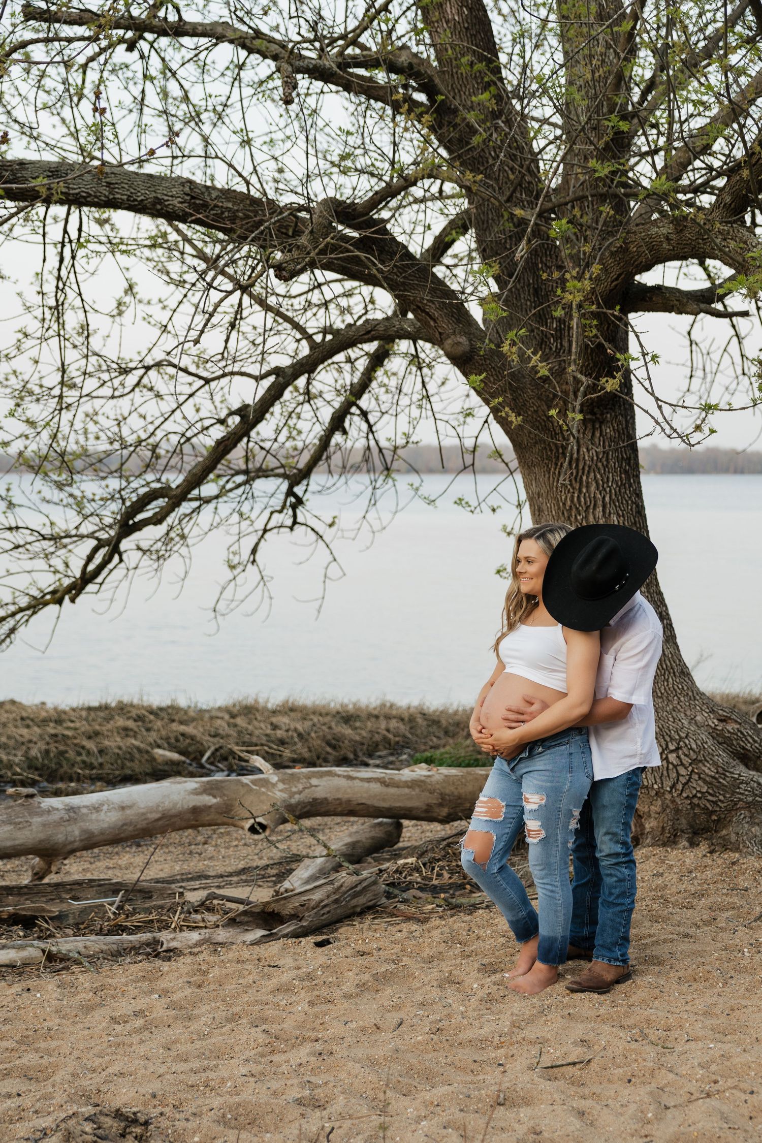 Expectant couple standing under lakeside tree, man in black cowboy hat hugging pregnant woman from behind.