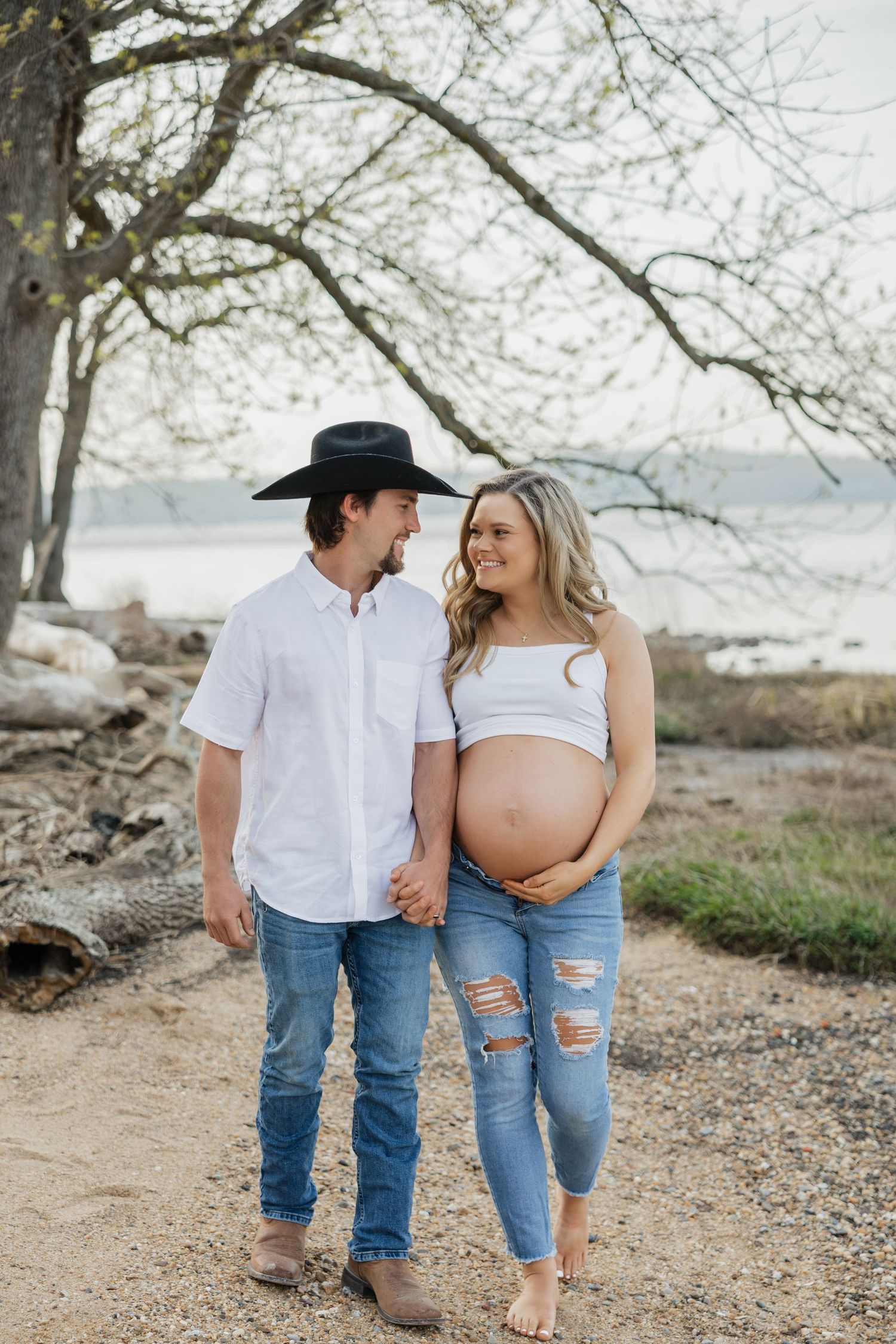 Couple sharing a tender moment on sandy path, pregnant woman in ripped jeans holding belly, man in cowboy hat.