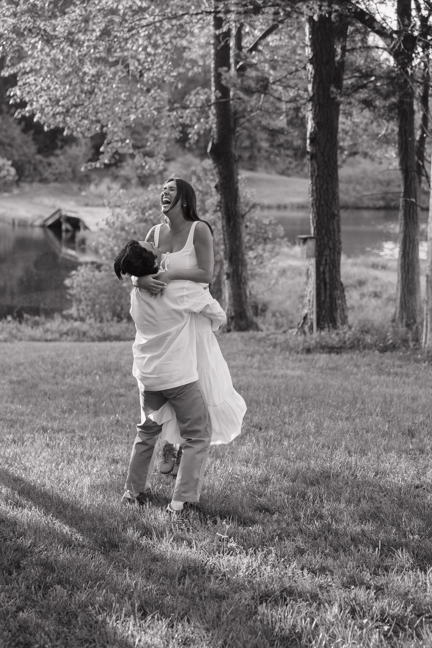Black and white photo of joyful couple, man lifting laughing woman in white dress in a grassy park.