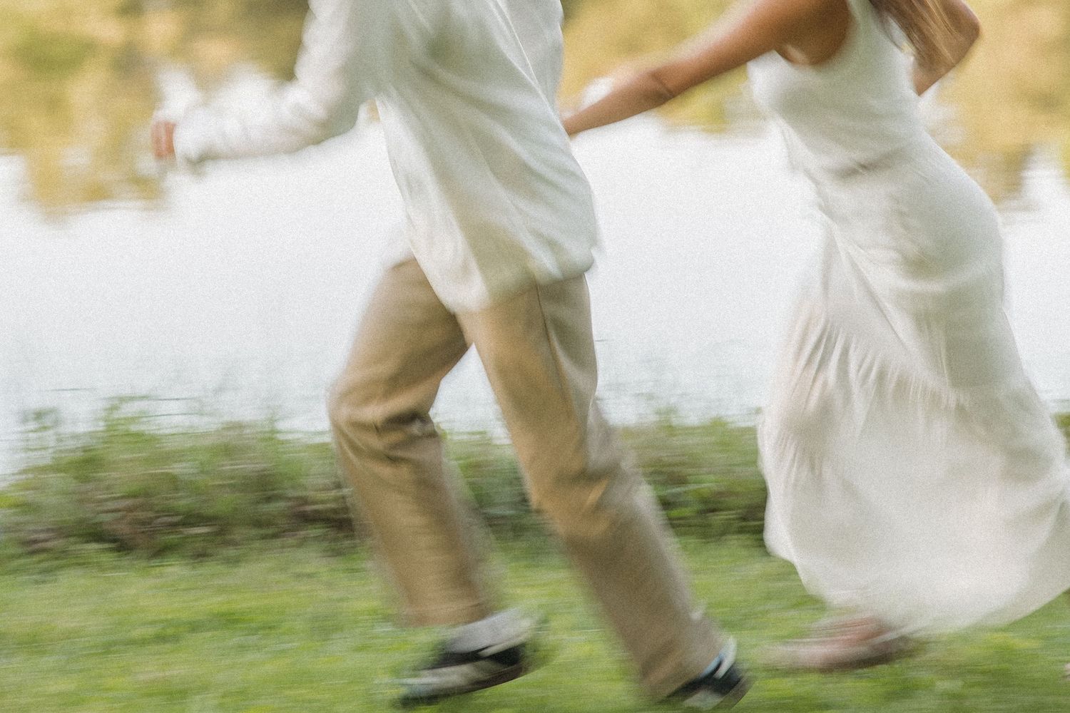 Motion-blurred couple running together in white outfits along a lush green lakeside in soft light.