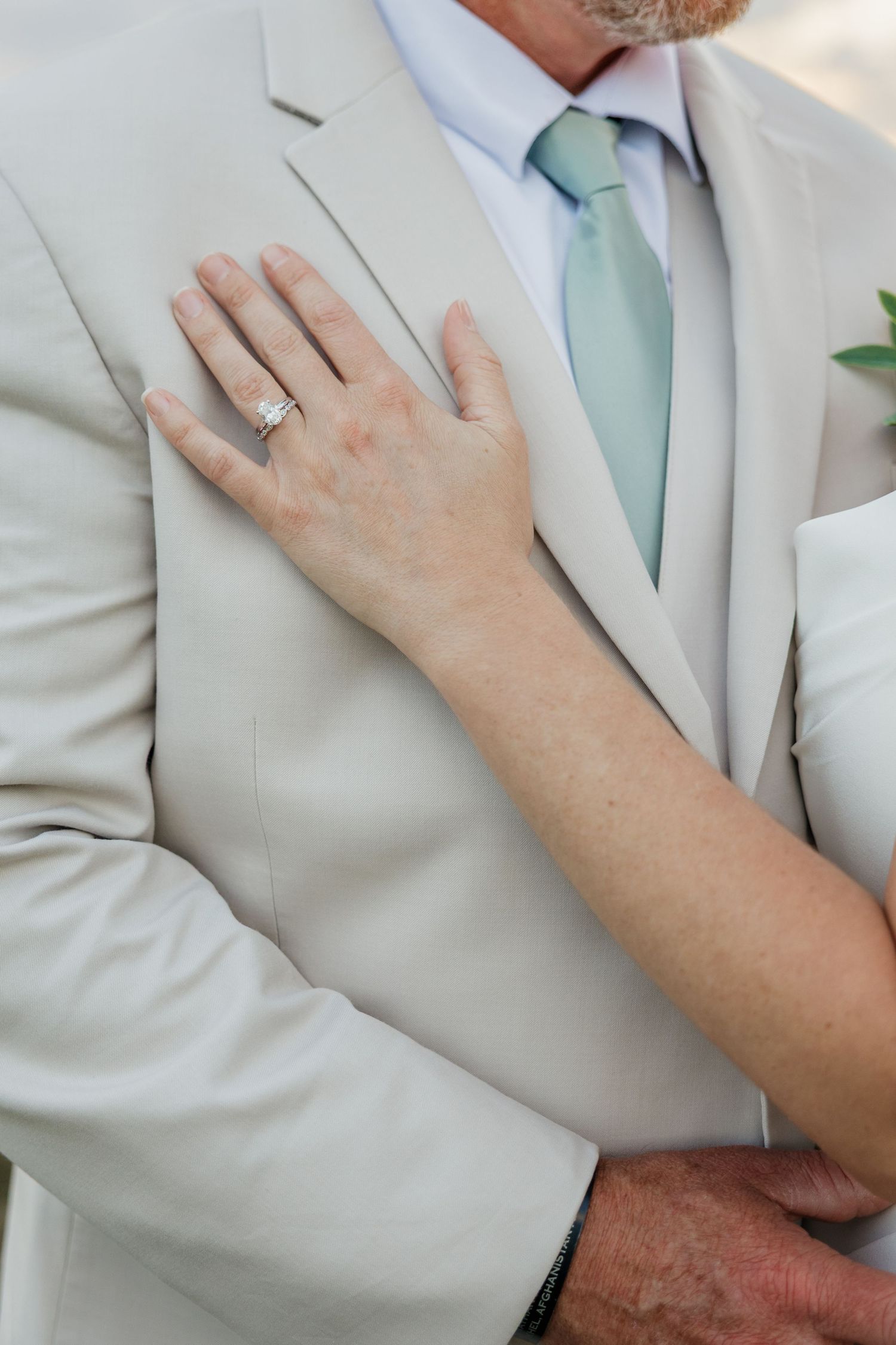 Close-up of bride's hand with diamond engagement ring resting on groom's beige suit with mint tie.