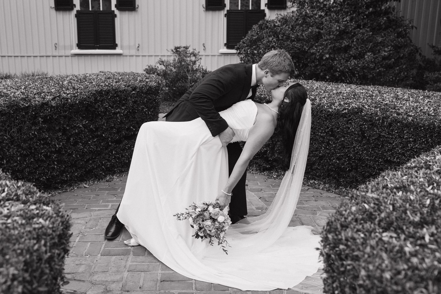 Black and white photo of bride and groom sharing a romantic dip kiss outside a chapel surrounded by hedges.