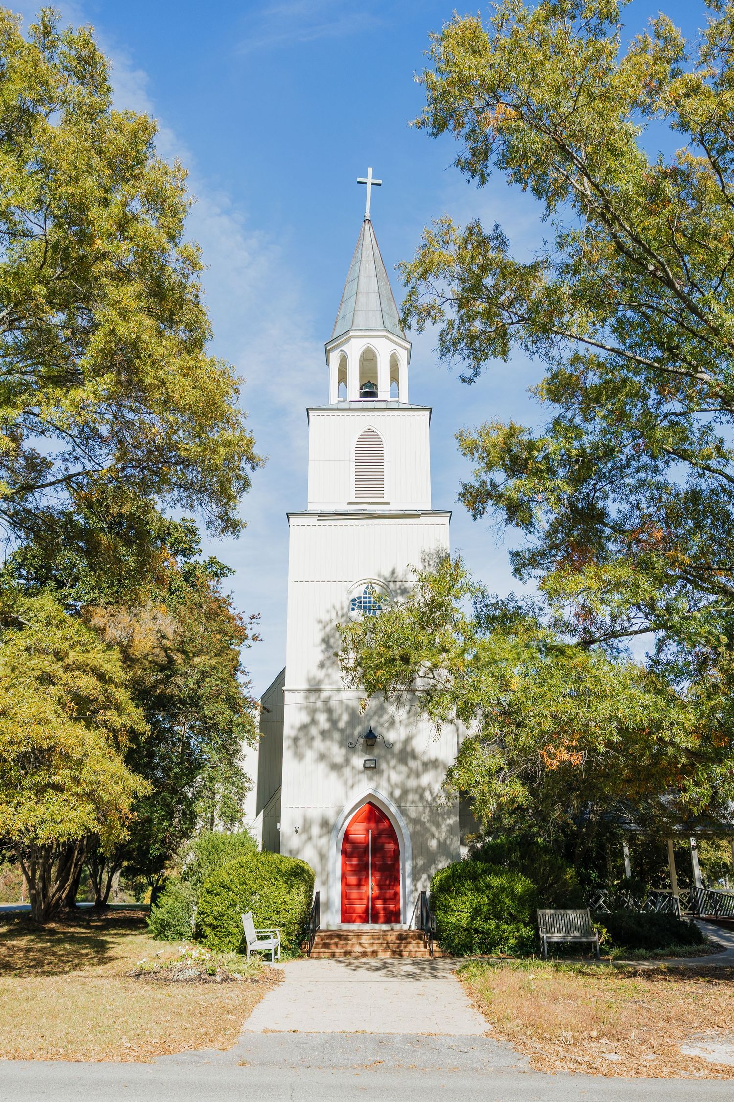 Classic white country church with red doors, steeple cross, and tall trees under a blue sky.