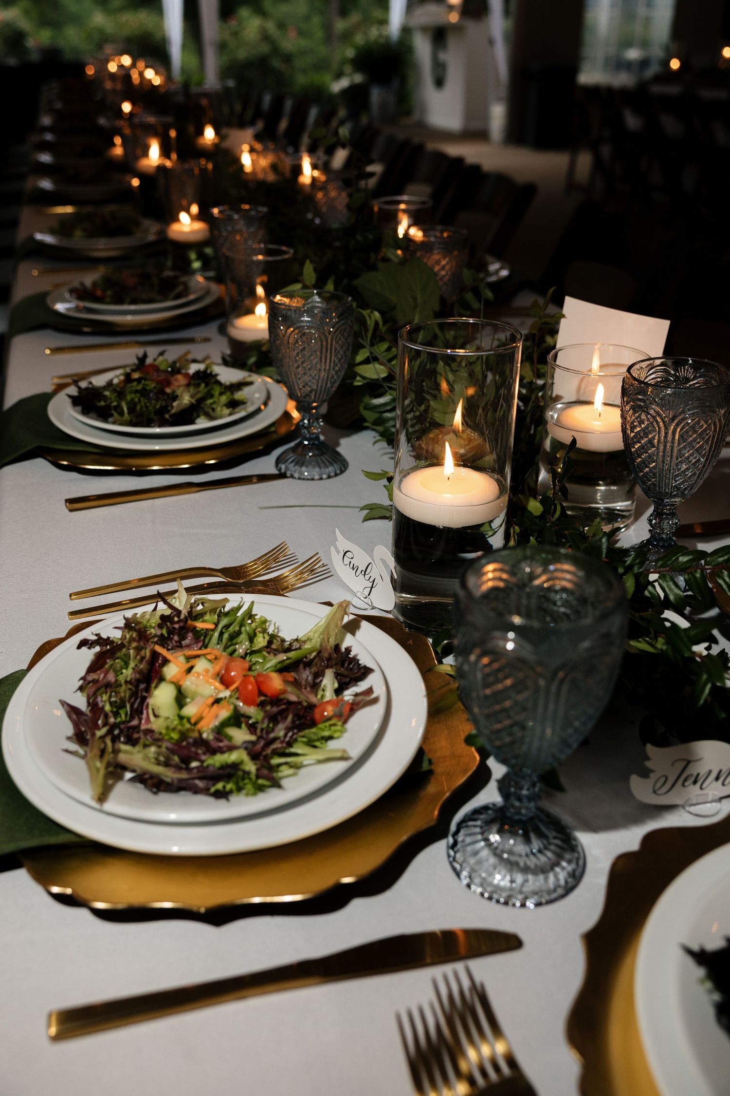 Elegant wedding reception table setting with candles, greenery, gold chargers, and fresh salads on white plates.