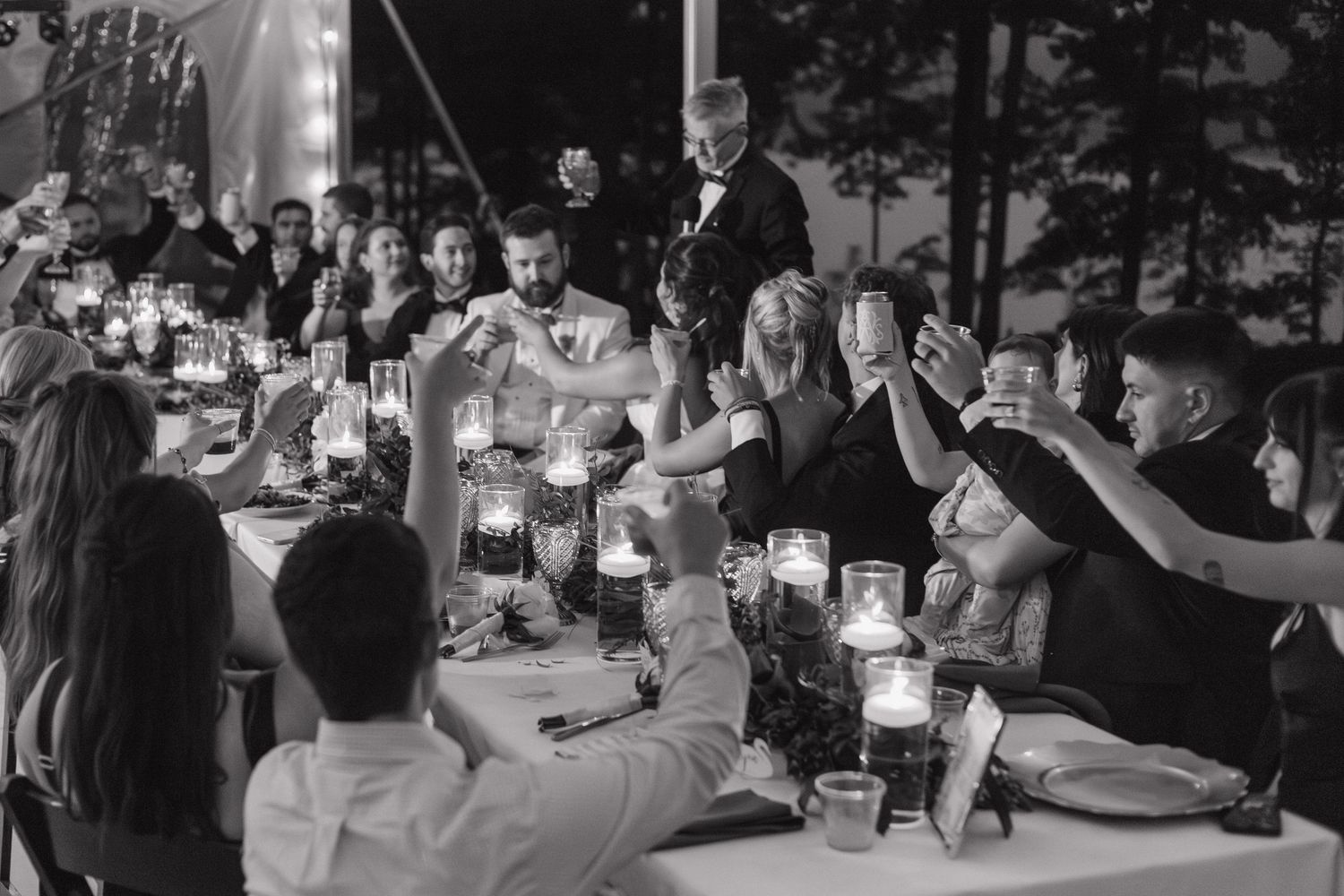 Black and white photo of wedding guests raising glasses in a toast at a candlelit reception dinner table.
