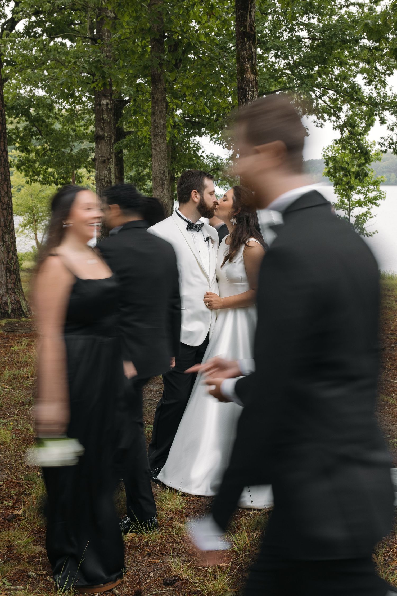 Artistic motion-blur photo of bride and groom kissing surrounded by moving wedding party guests in forest.