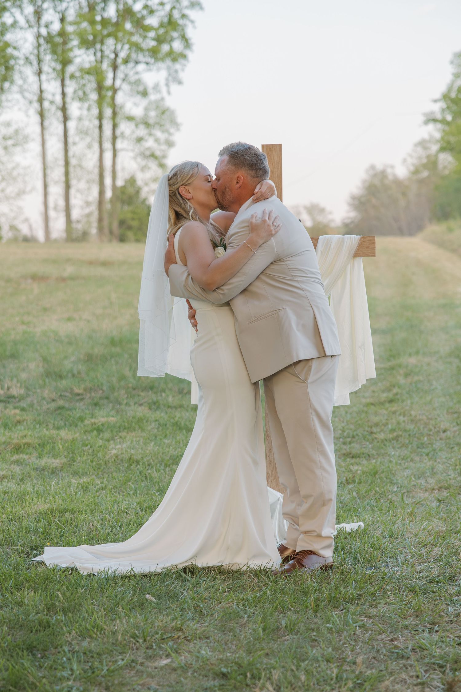 Bride and groom share their first kiss at an outdoor wedding ceremony in front of a wooden cross.