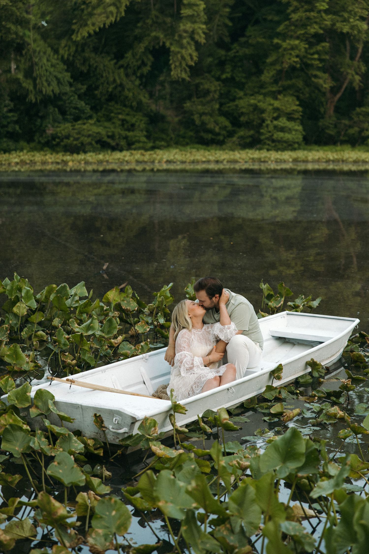 A couple shares a romantic kiss while sitting together in a white rowboat surrounded by green lily pads.
