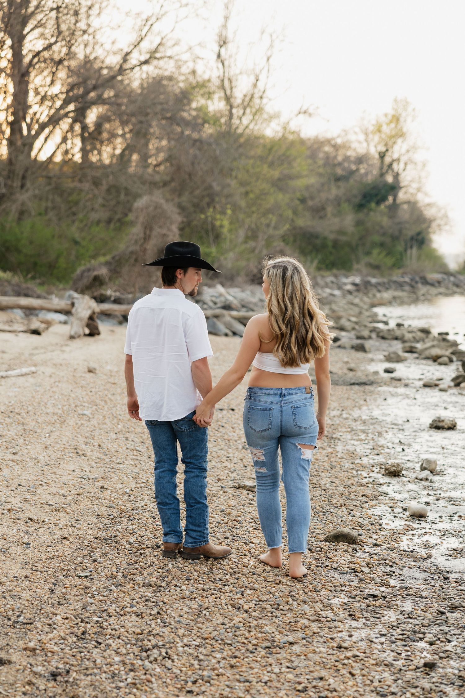 Couple holding hands walking along rocky riverbank, man wearing black cowboy hat and white shirt.