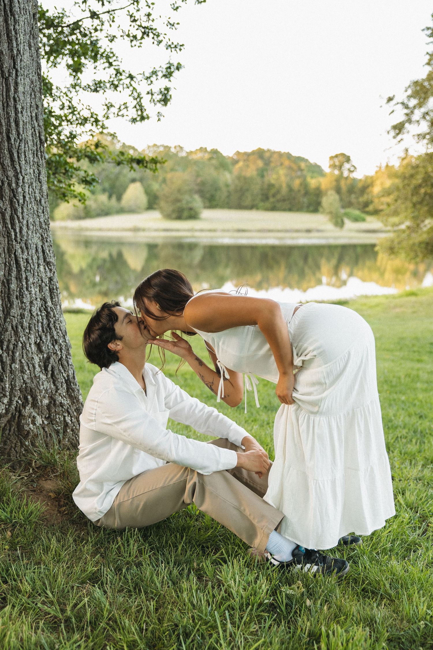 A romantic couple shares a kiss near a tree beside a peaceful reflective lake at sunset.