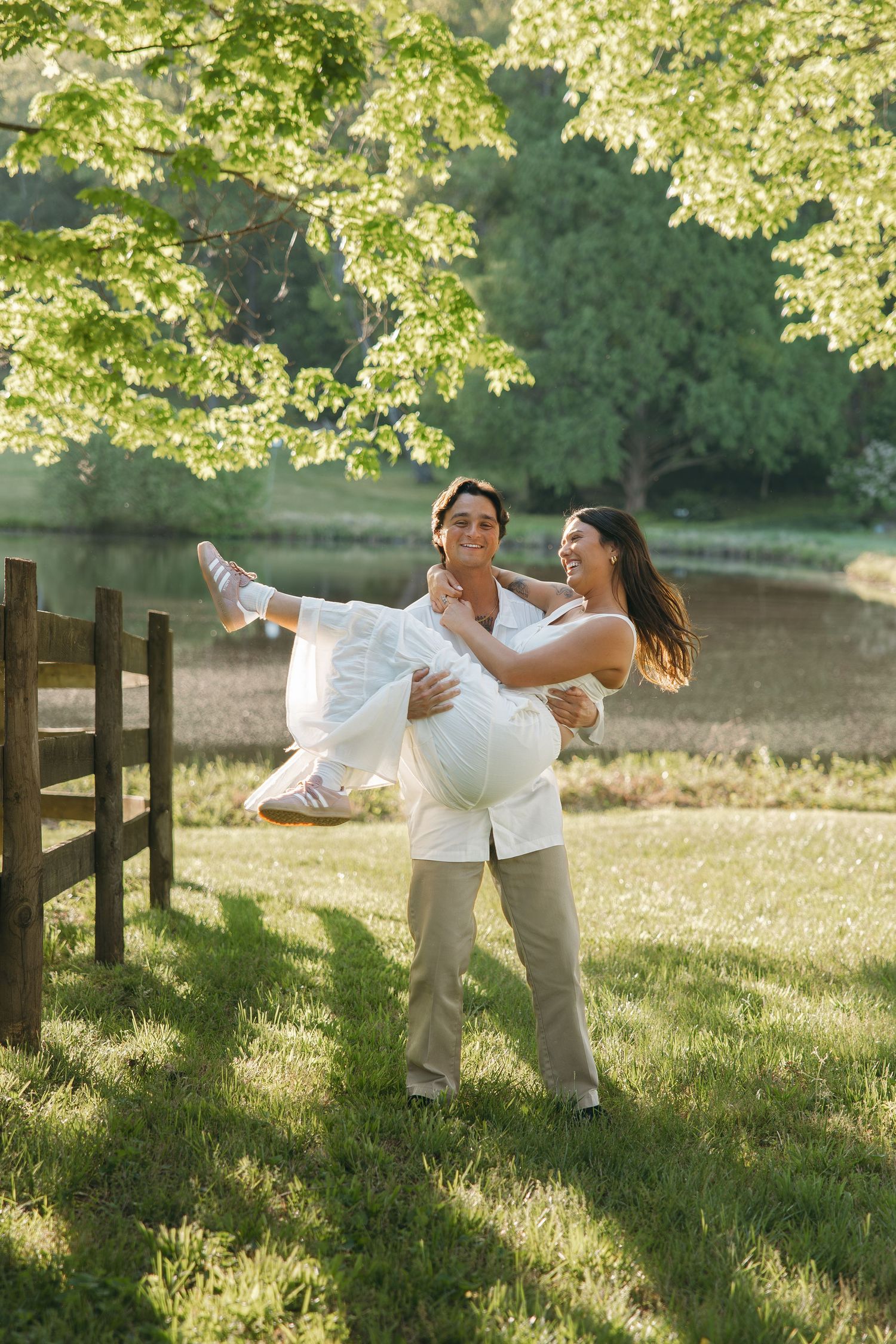 Happy couple laughing as man carries woman in white dress in a sunlit green meadow by a lake.