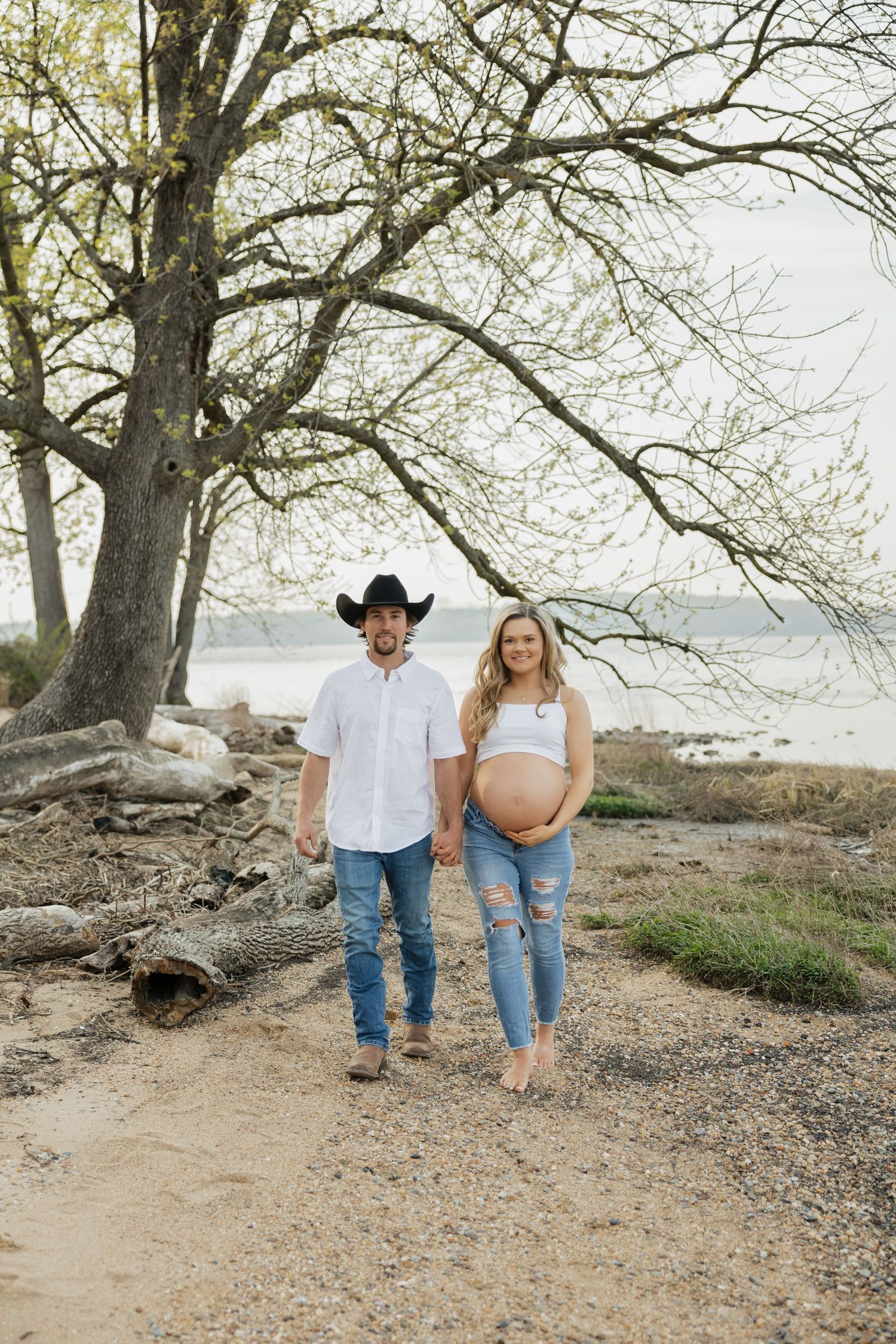 Smiling pregnant couple holding hands on sandy path near lake, man in black cowboy hat and white shirt.