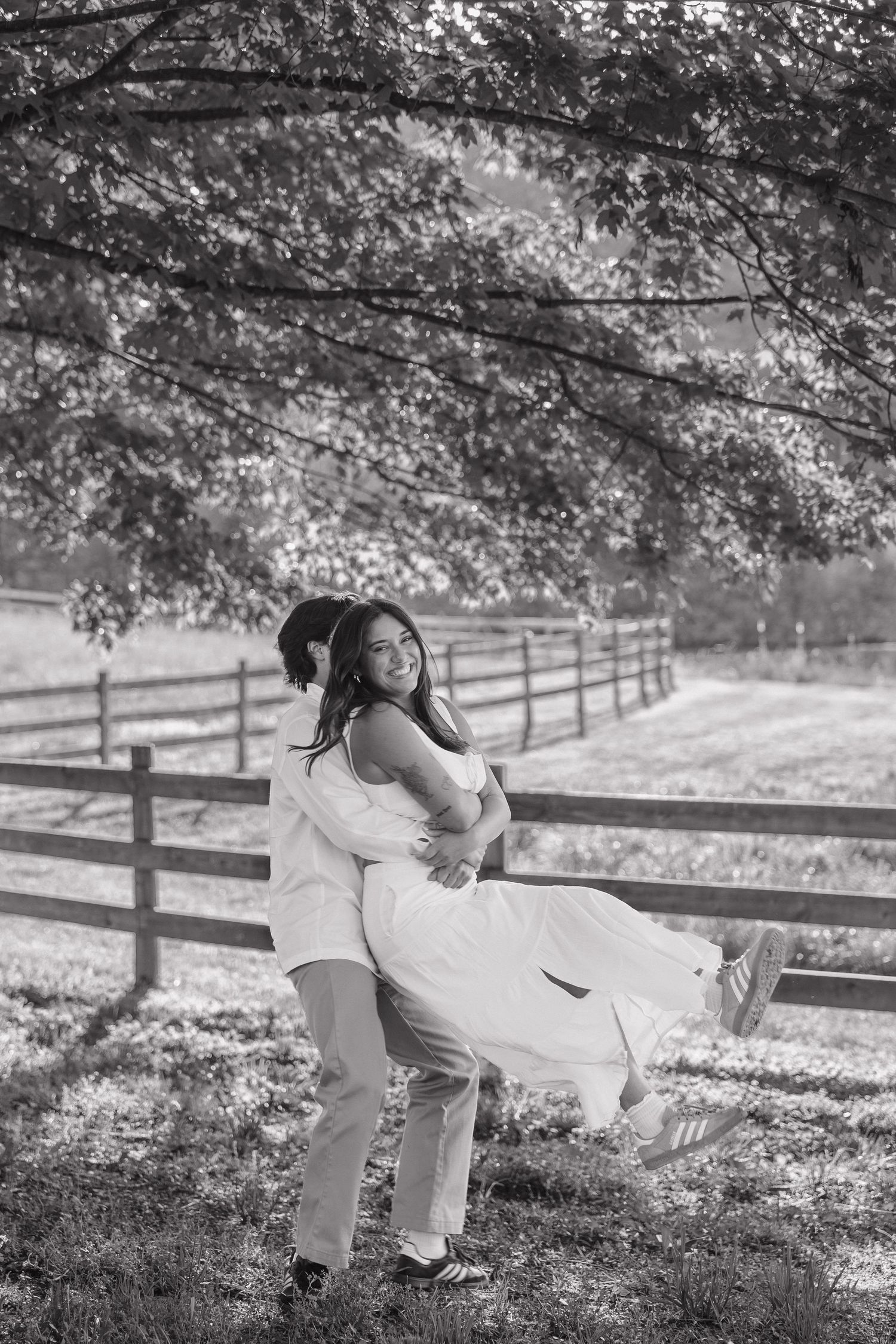 Black and white photo of groom lifting smiling bride under a canopy of trees near a wooden fence.