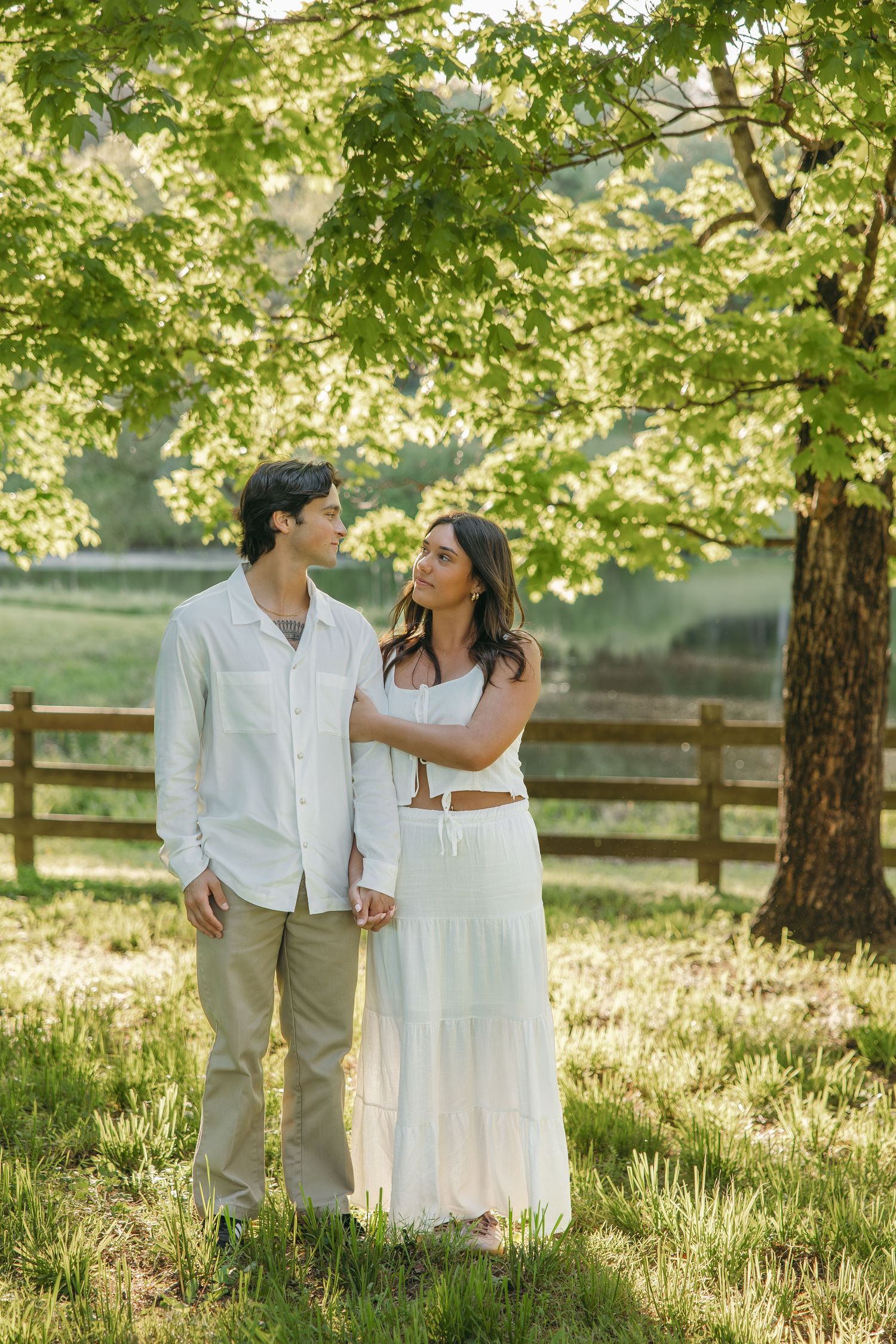 Couple holding hands and gazing at each other under a large tree in a sunny green meadow.