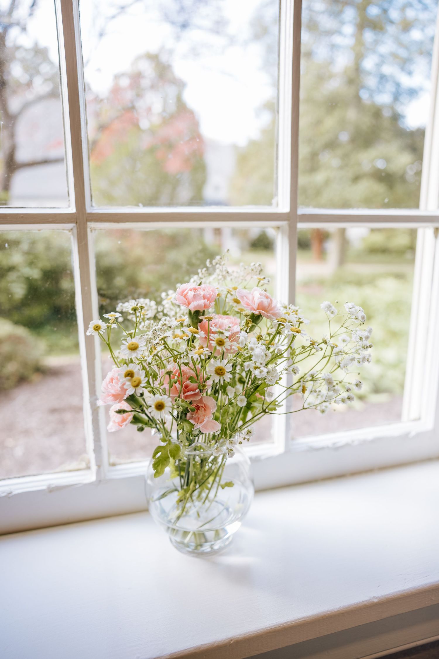 A glass vase of pink roses, daisies, and wildflowers sits on a white windowsill.