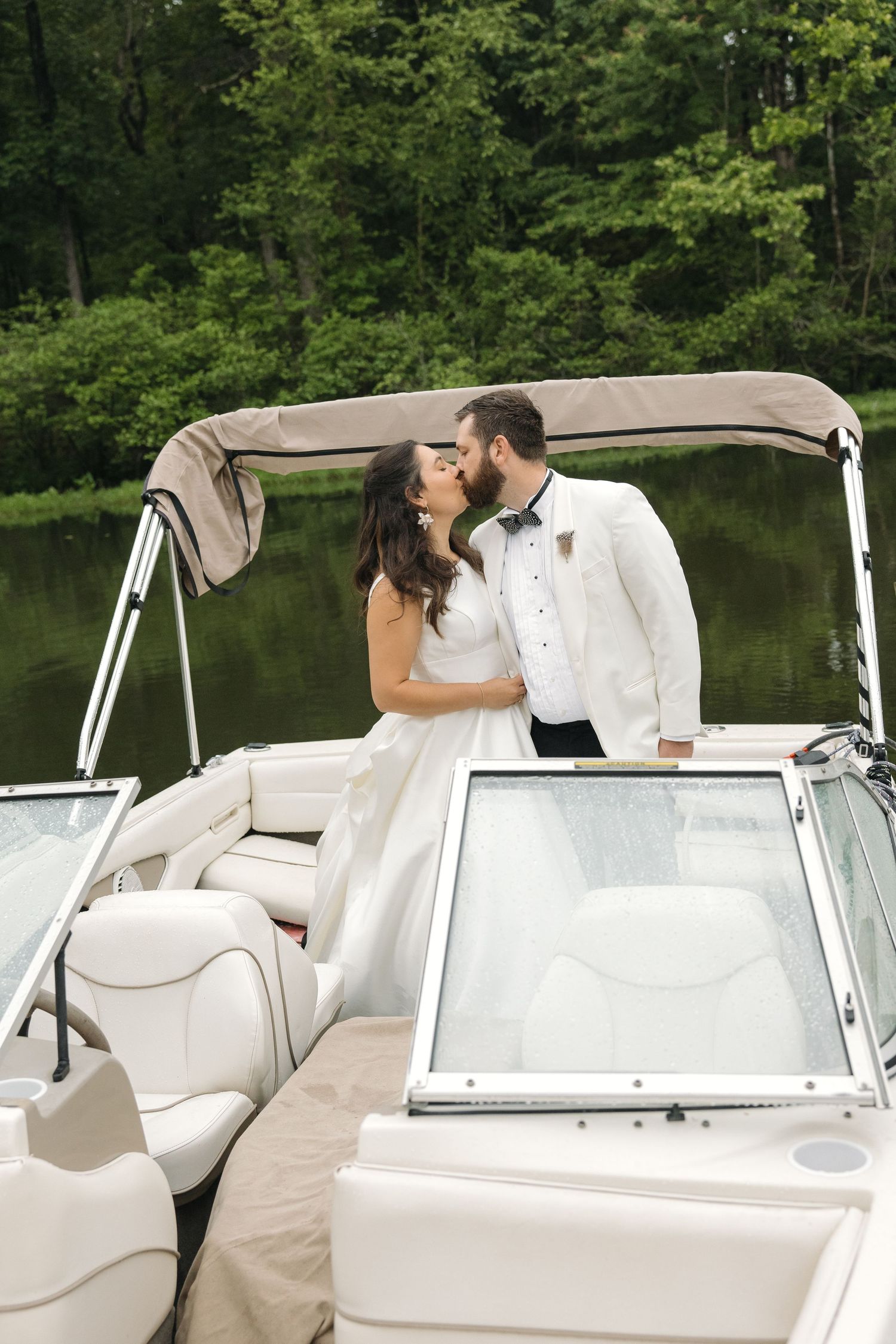 Romantic bride and groom sharing a kiss on a white luxury boat with green forest backdrop.