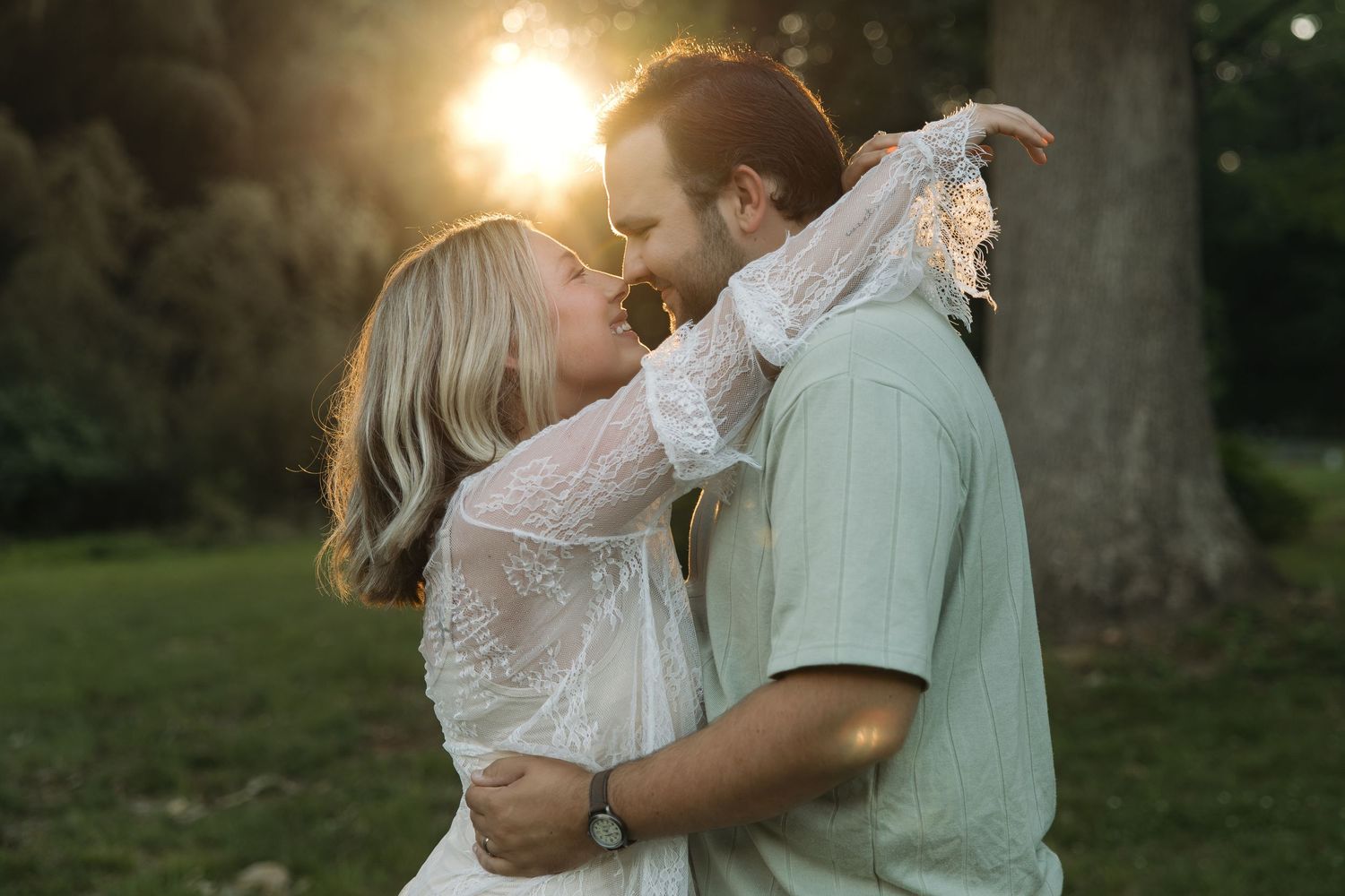 Romantic couple sharing a kiss backlit by warm golden sunset glow among green trees outdoors.