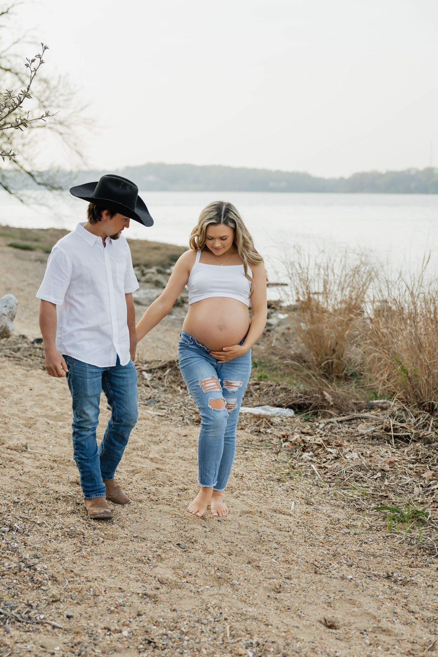 Expectant couple strolls along sandy lakeside beach, woman cradling baby bump while man in cowboy hat looks on.