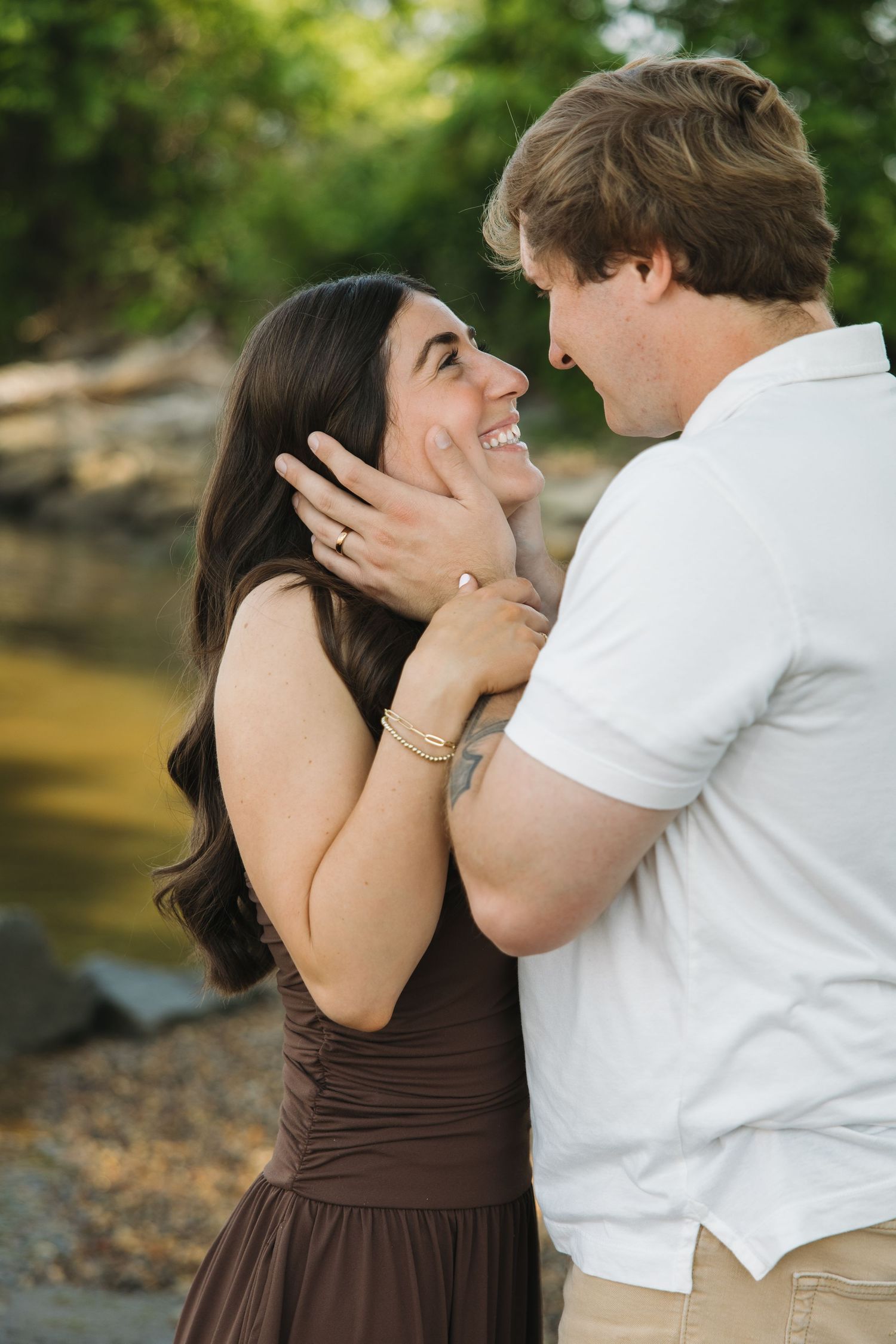 Close-up of a smiling couple gazing at each other near a creek, woman wearing engagement ring.