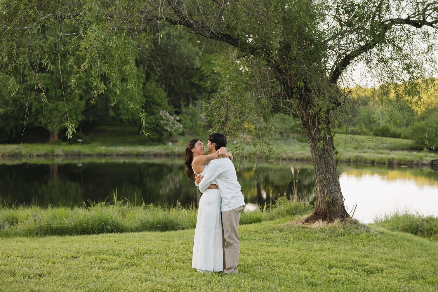 Couple embracing and kissing in a green park with a peaceful pond and willow tree backdrop.