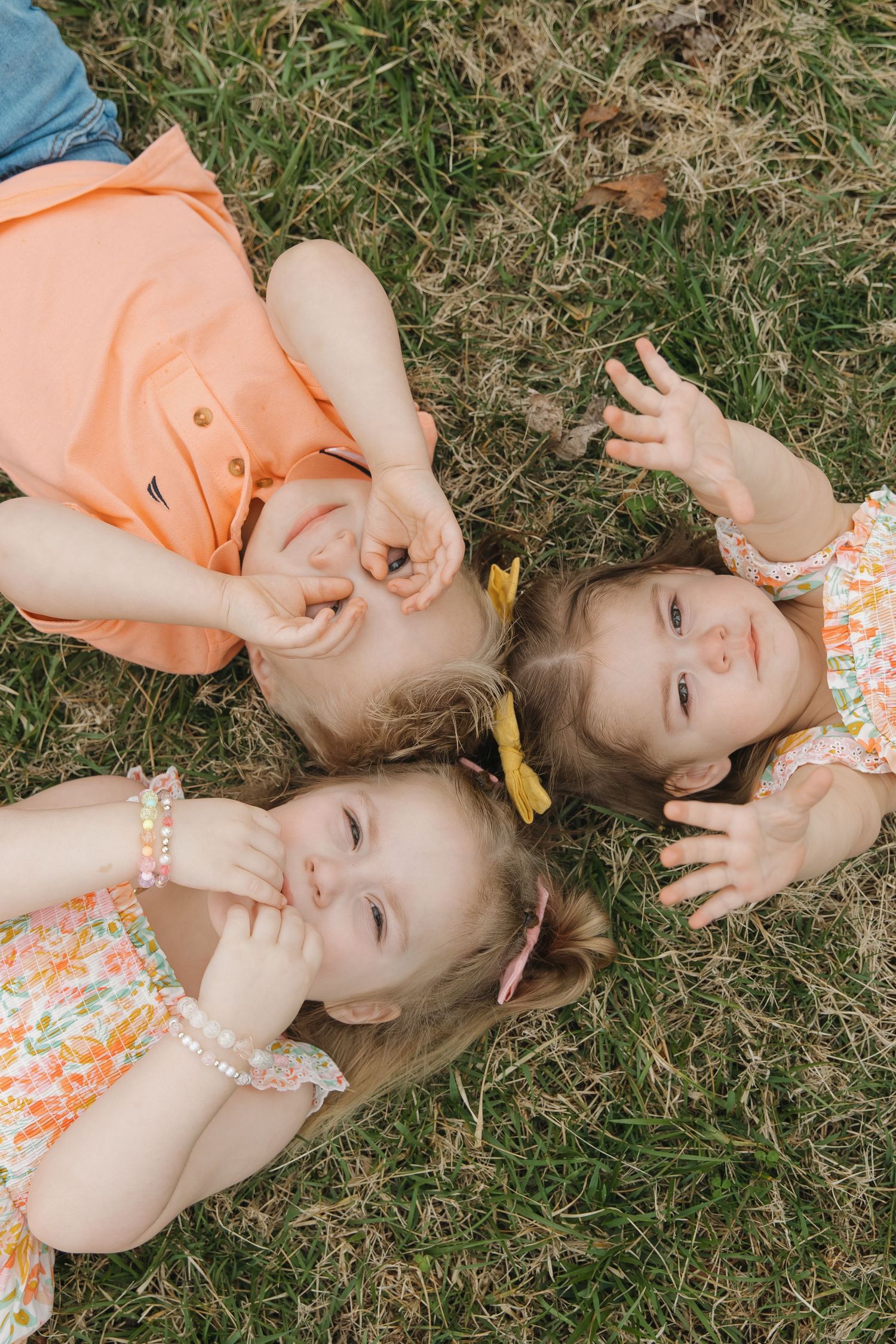 Three young children in orange and floral outfits lie on grass looking up at the camera with playful expressions.