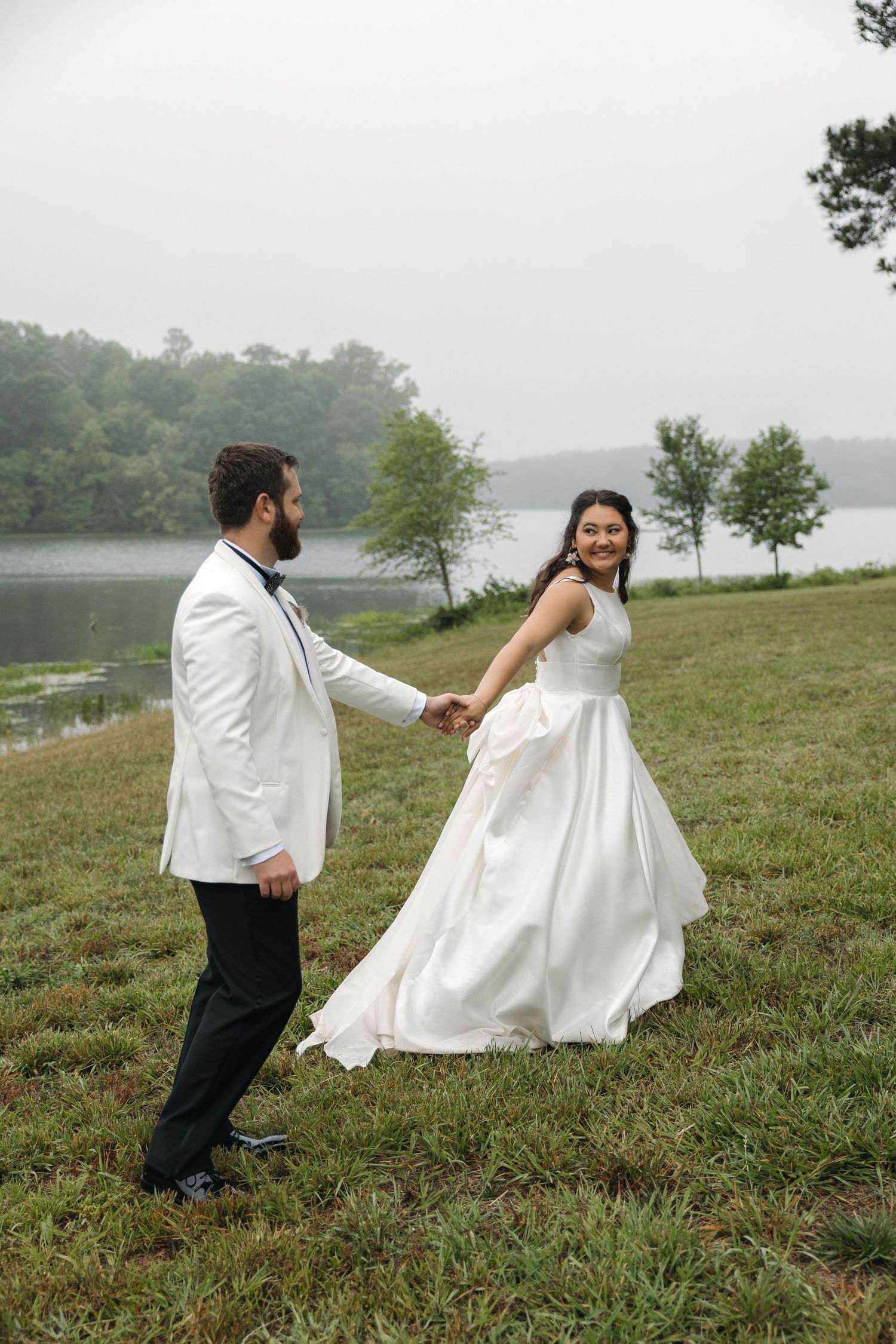 Smiling bride leads groom by hand across green lawn with foggy lake behind.