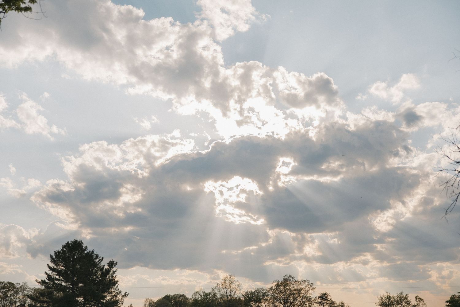 Dramatic sunrays breaking through fluffy clouds over treetops in bright sky.