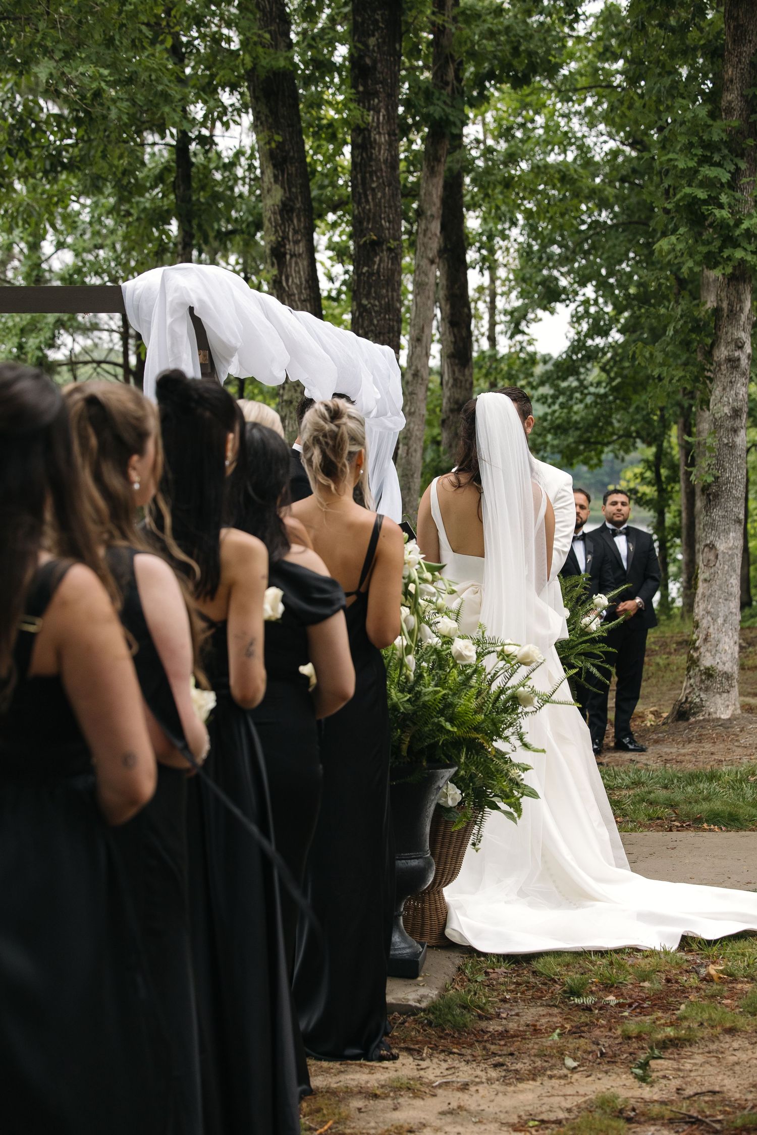 Bridesmaids in black gowns line the aisle as bride in white dress with veil walks toward groom at outdoor wedding.