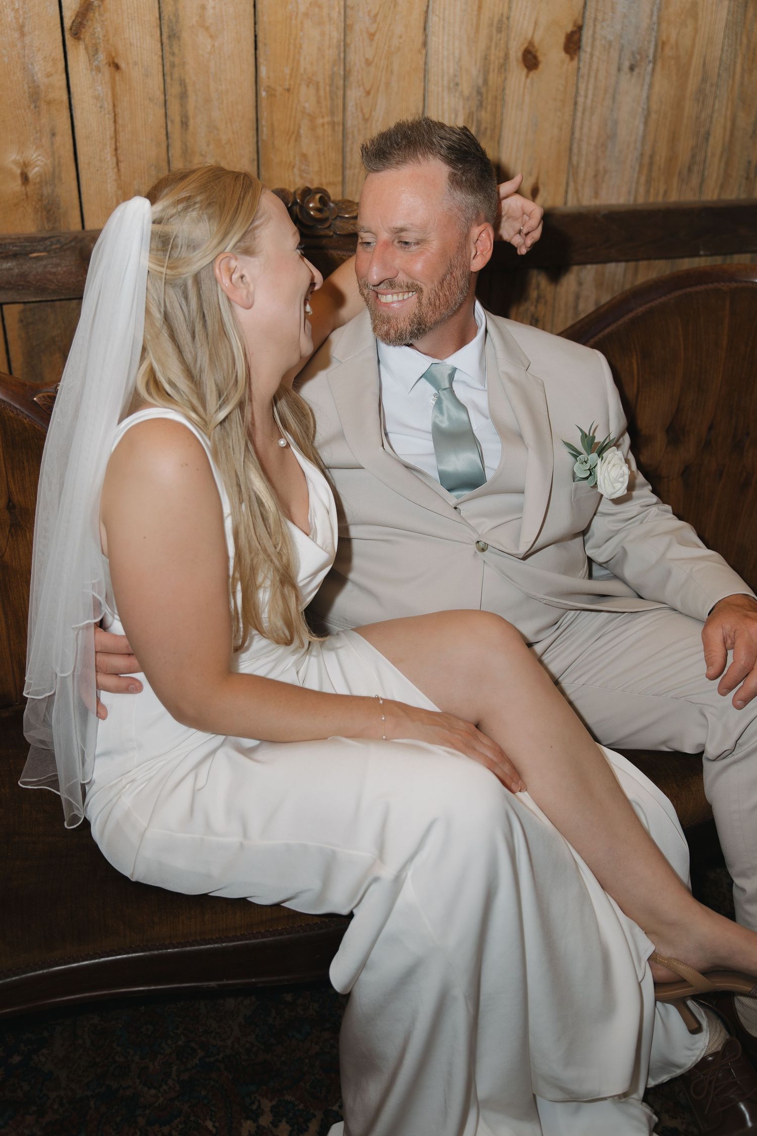 Bride sitting on grooms lap laughing together on a wooden bench inside a rustic barn.