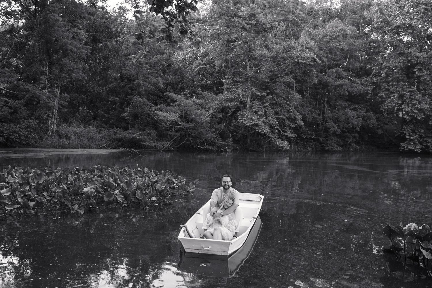 Black and white photo of a person sitting alone in a white boat on a peaceful river.