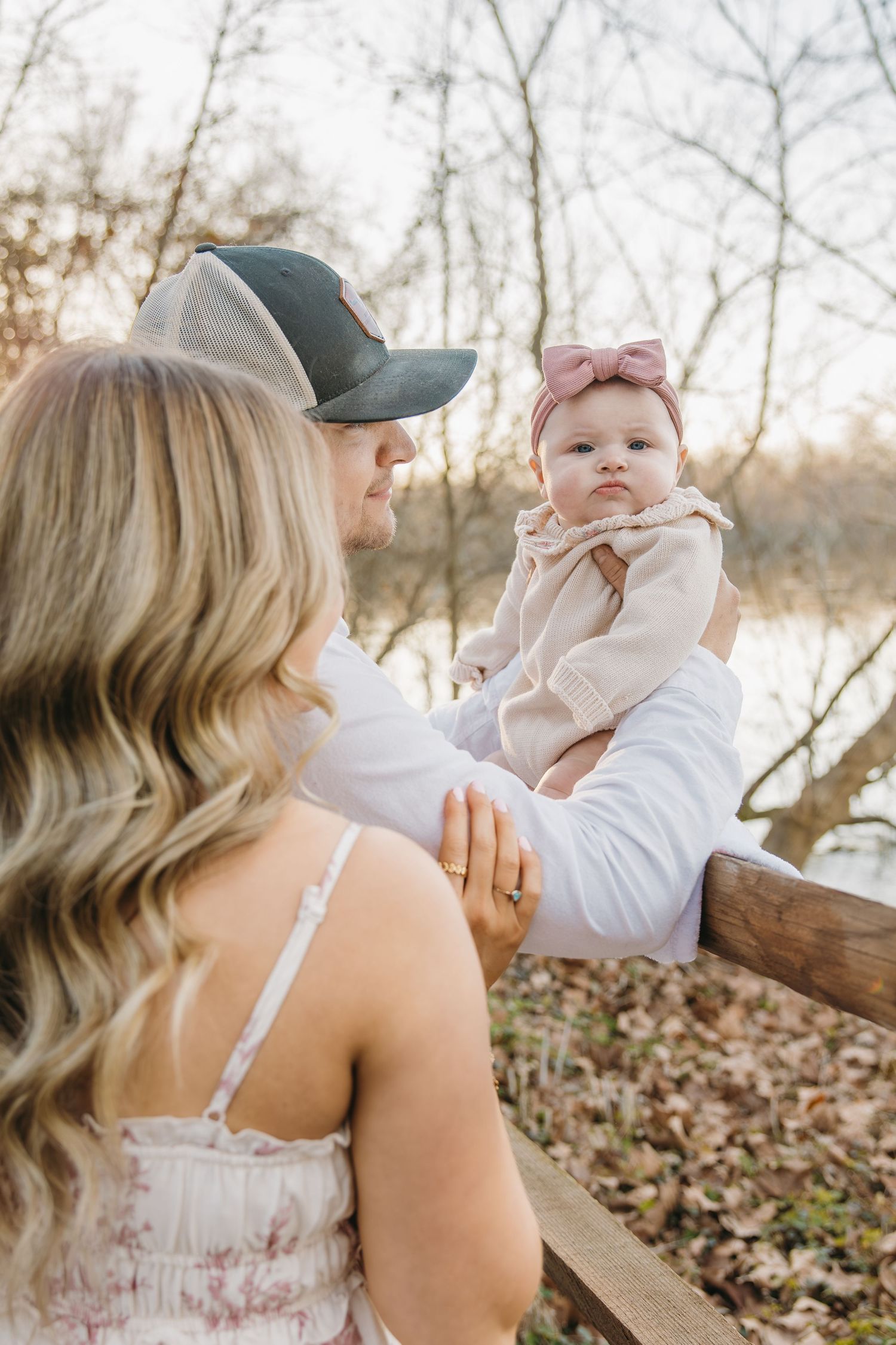Parents lovingly gazing at their baby girl with pink bow headband near a wooden fence outdoors.
