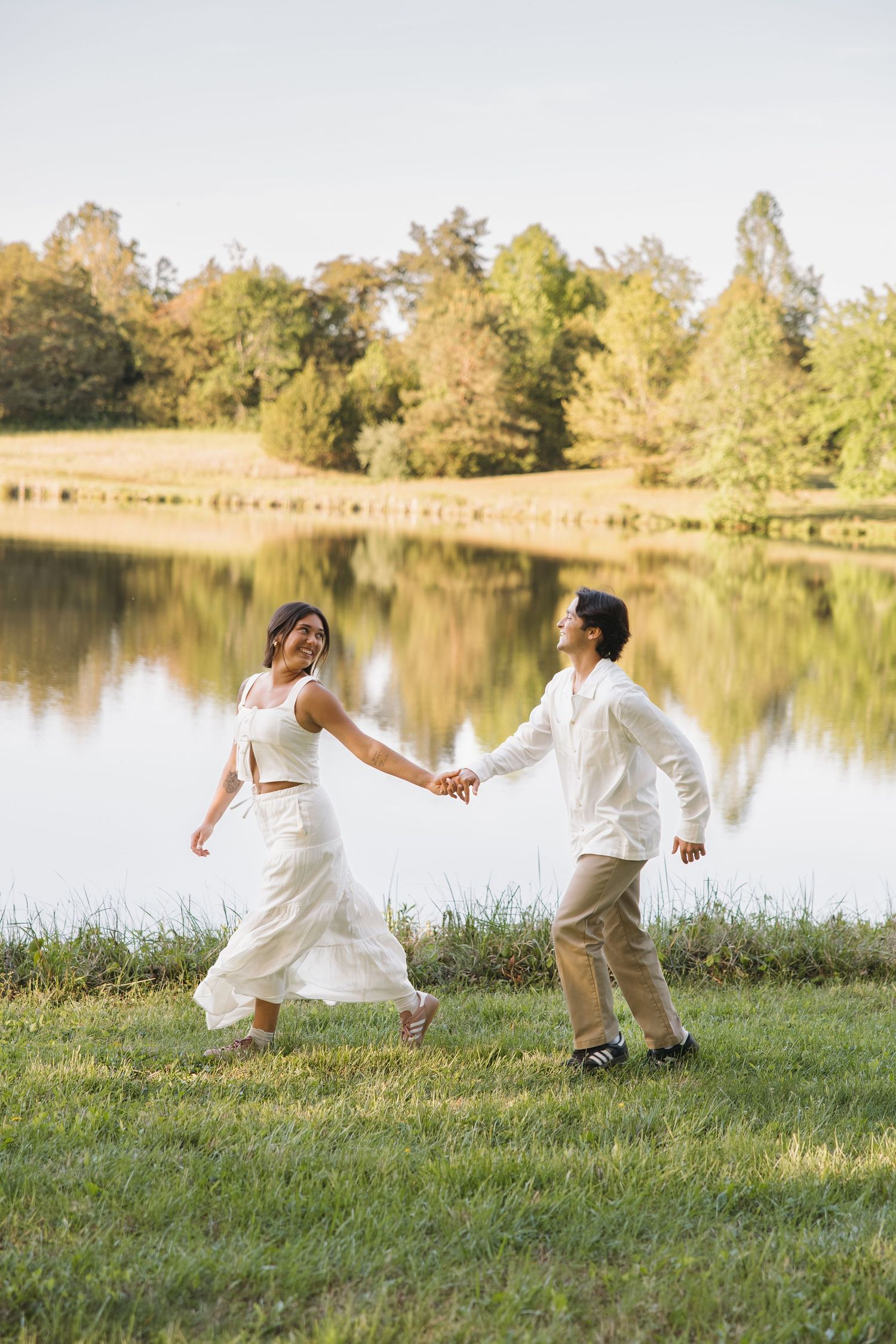 Laughing couple in white outfits holding hands and walking along a reflective lake surrounded by autumn trees.