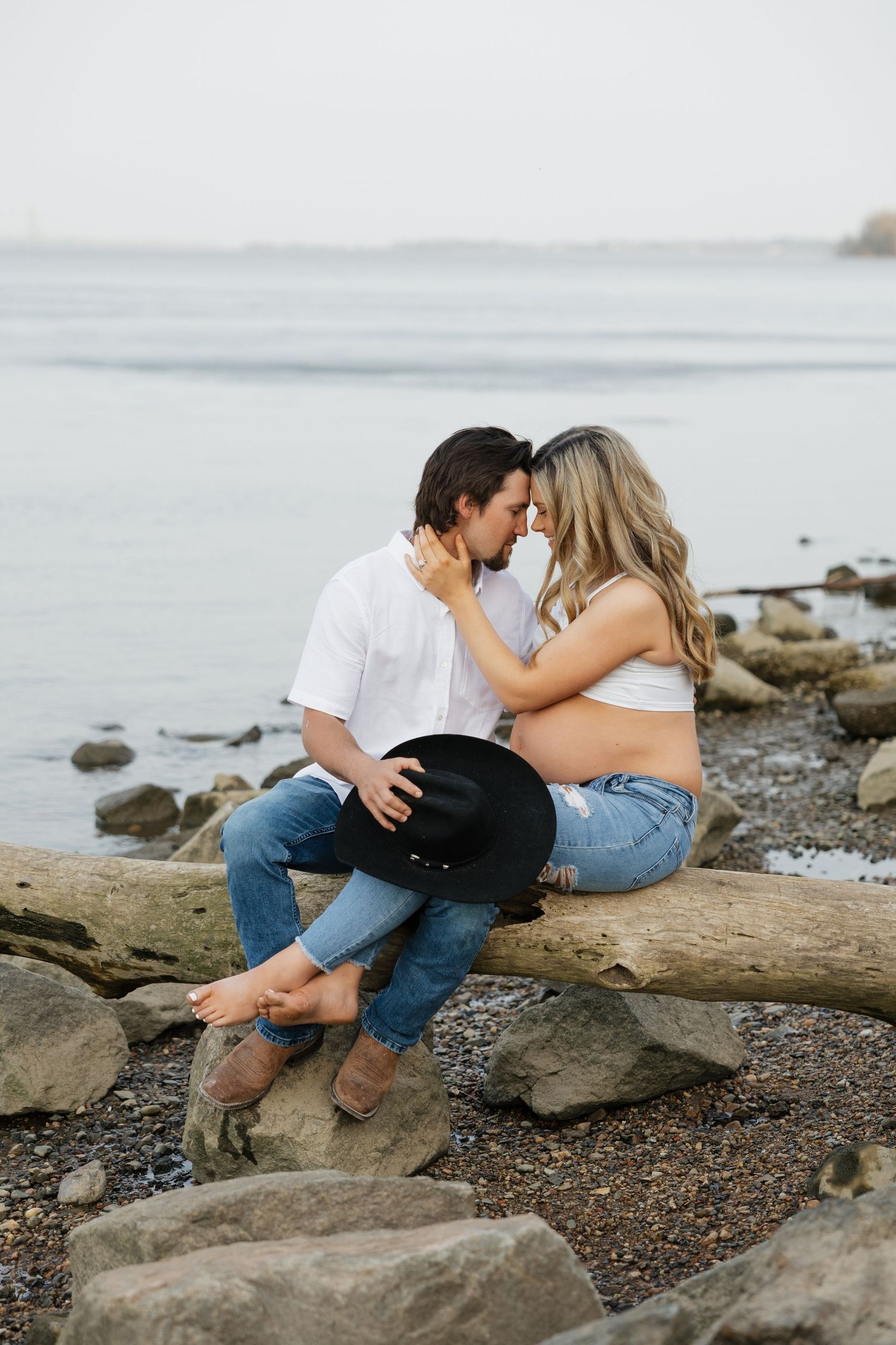 Couple sharing a tender nose-to-nose moment on driftwood by the lake, man holding black cowboy hat.