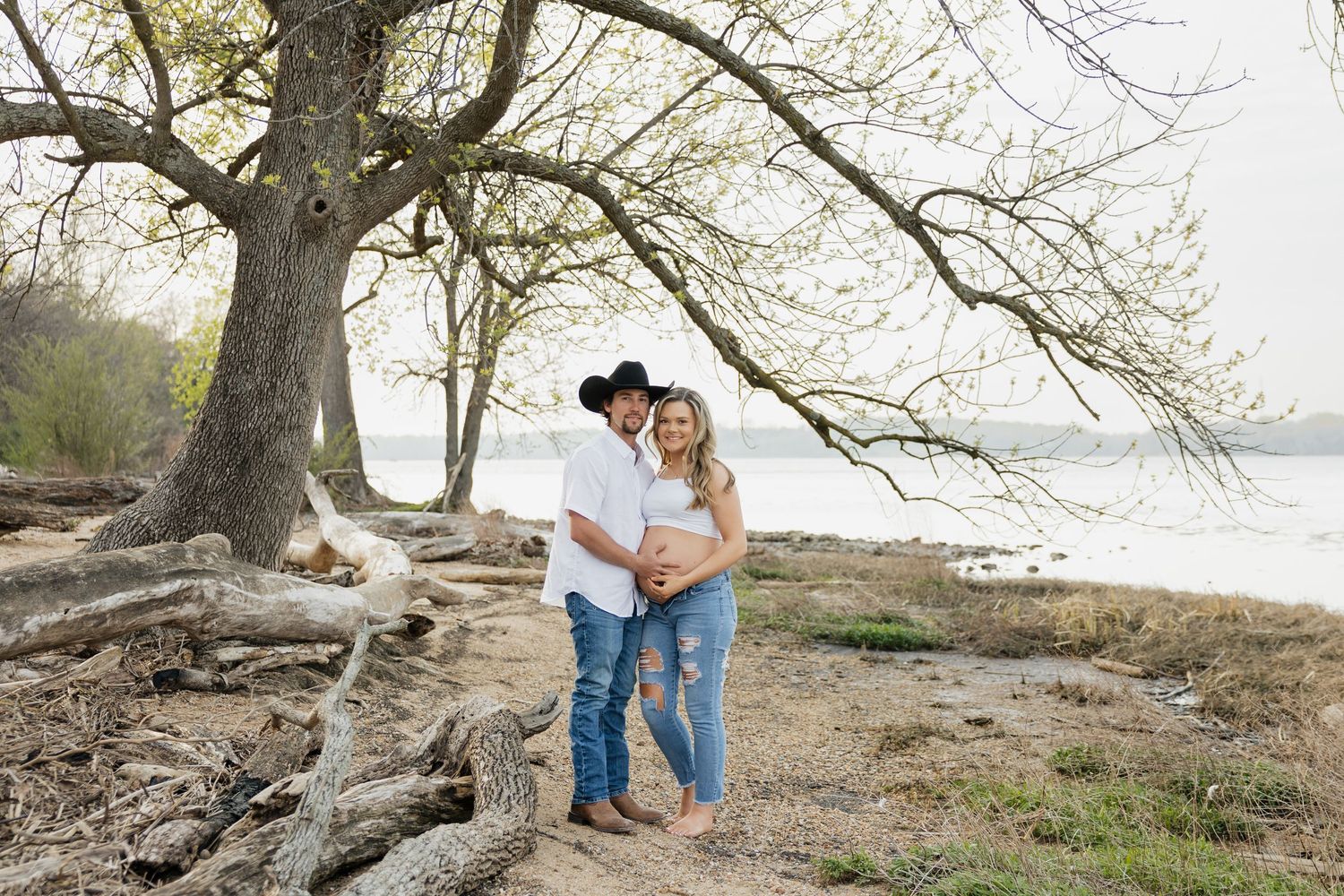 Expecting couple embracing under a large tree by a lake, man in cowboy hat with hands on baby bump.