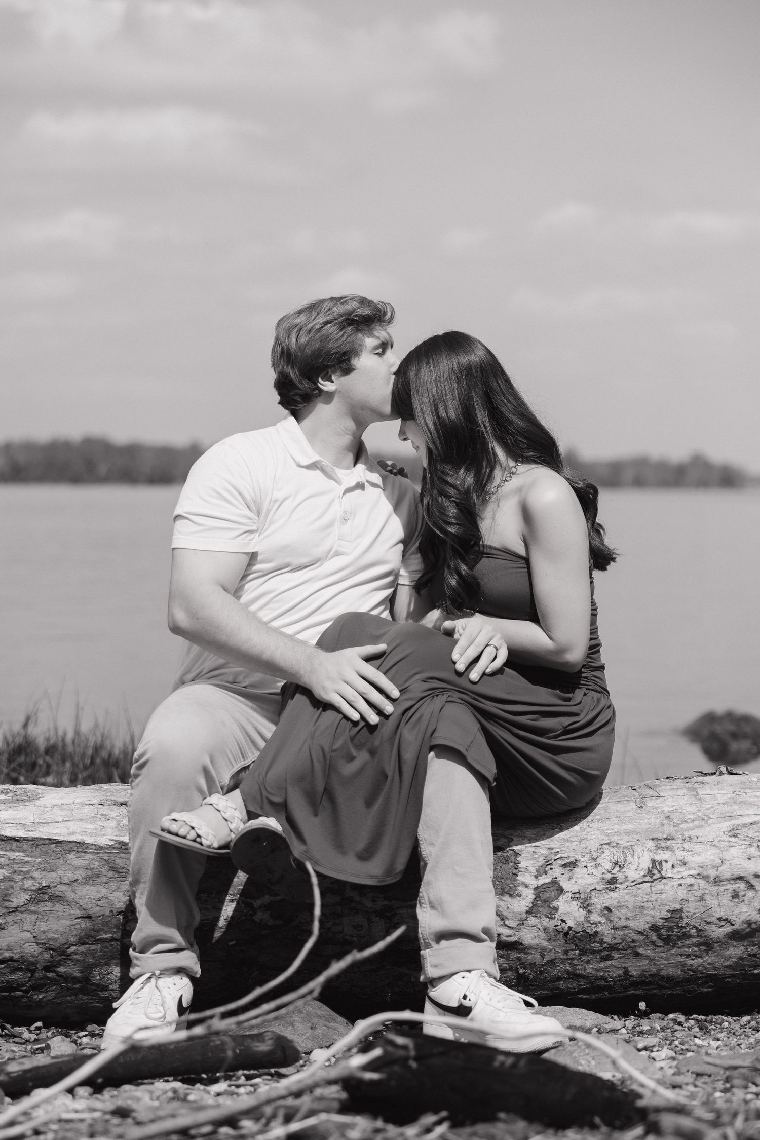 Black and white photo of a couple sharing a kiss while sitting on a log by a serene lakeside.